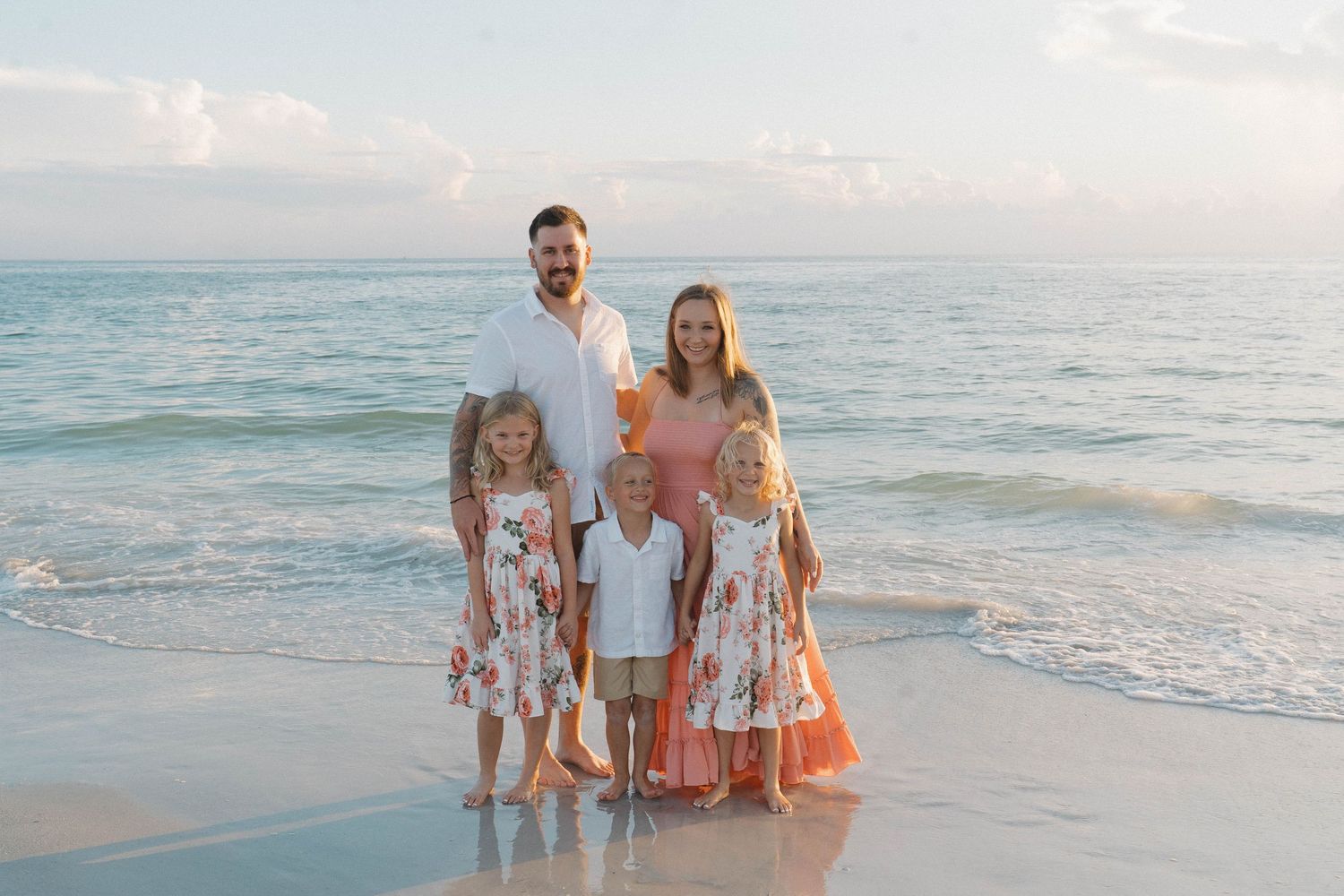 Family of five posing on a sandy beach at sunset, with ocean waves and pastel sky in the background.