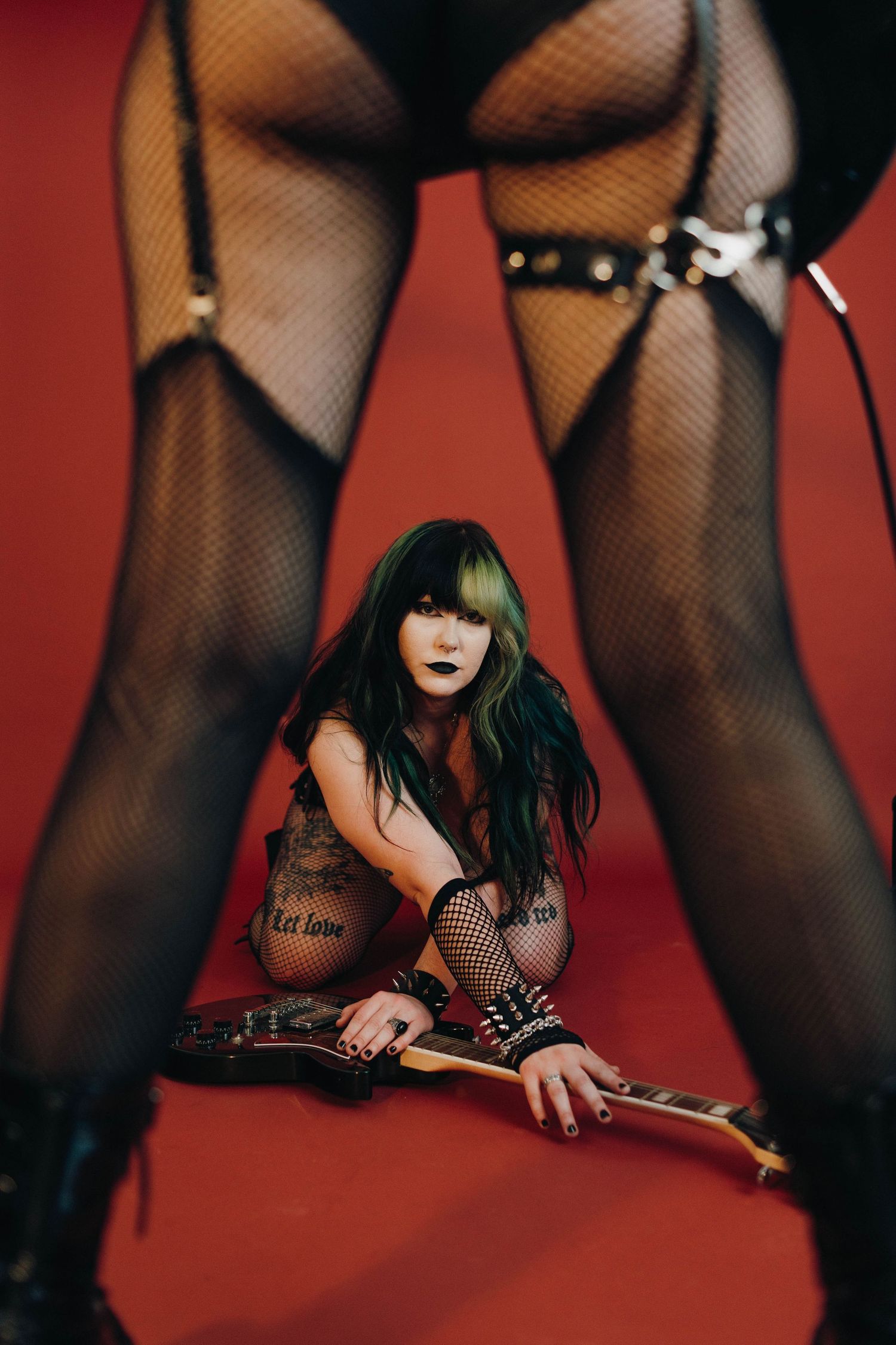 Punk rock woman with green hair sits on red floor holding electric guitar wearing fishnet stockings and leather accessories.