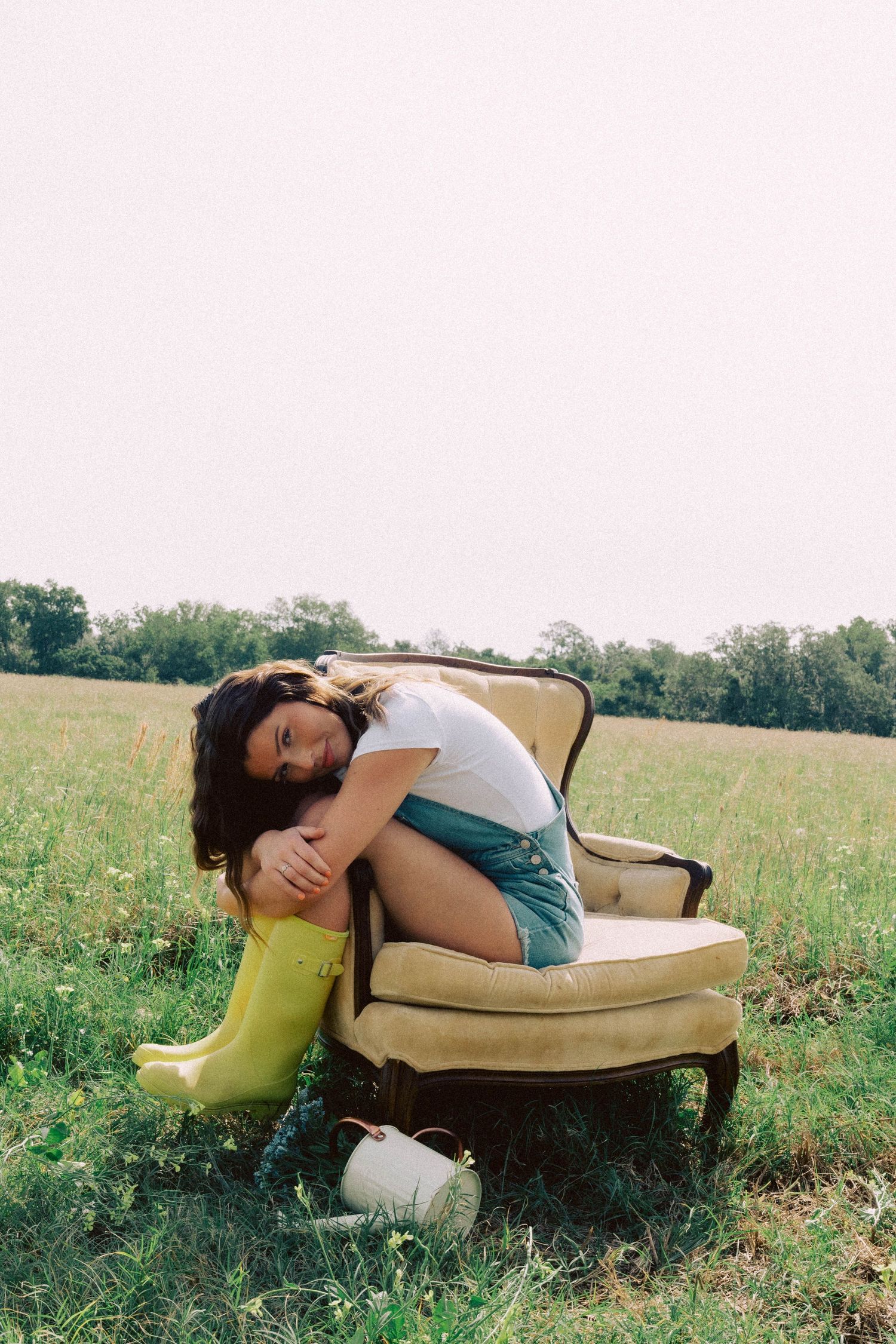 Woman in white tee, denim shorts, and yellow rain boots lounging on vintage armchair in a green field.