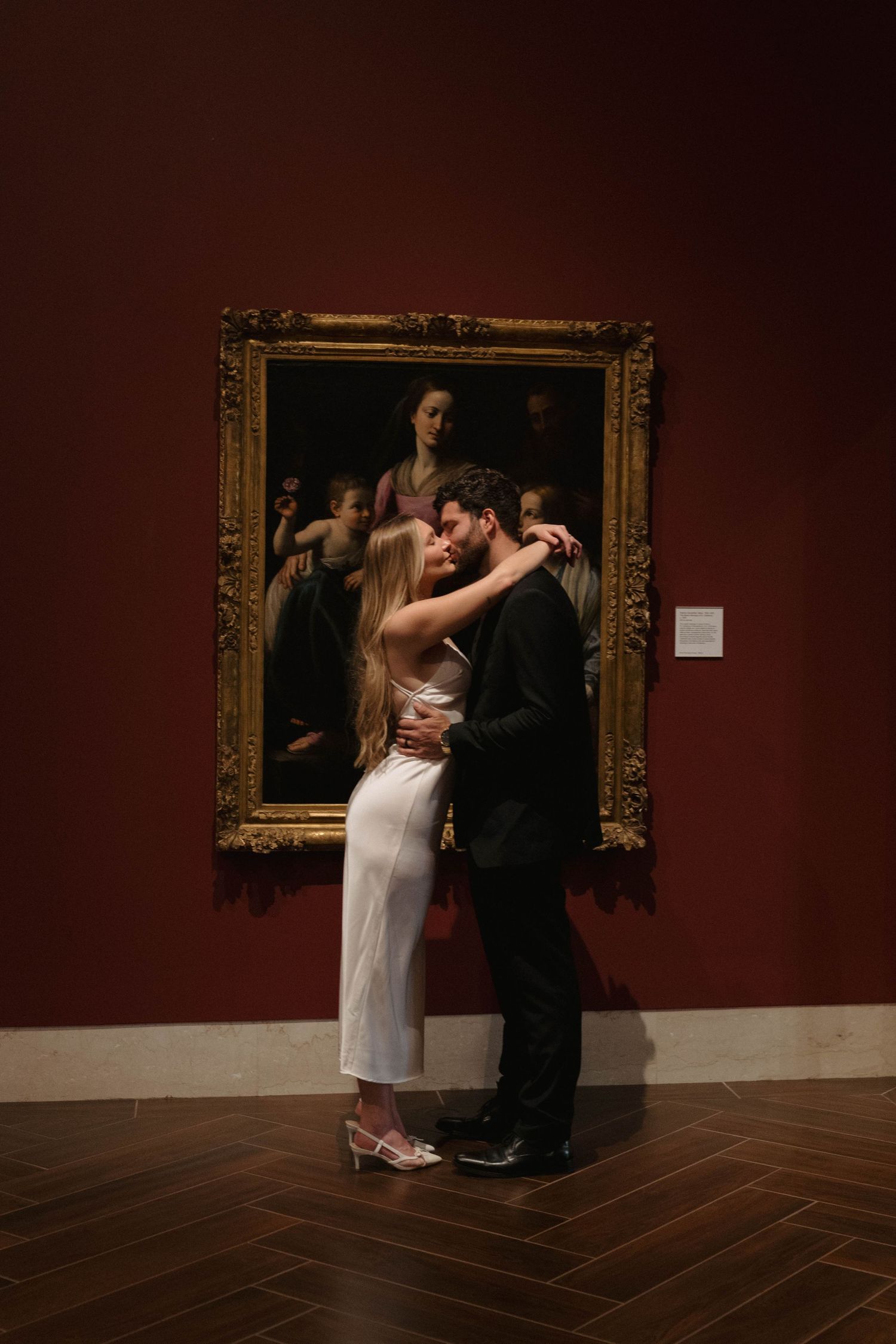 A couple embraces in front of an ornate gold-framed painting in a dimly lit museum with burgundy walls and wooden floors.
