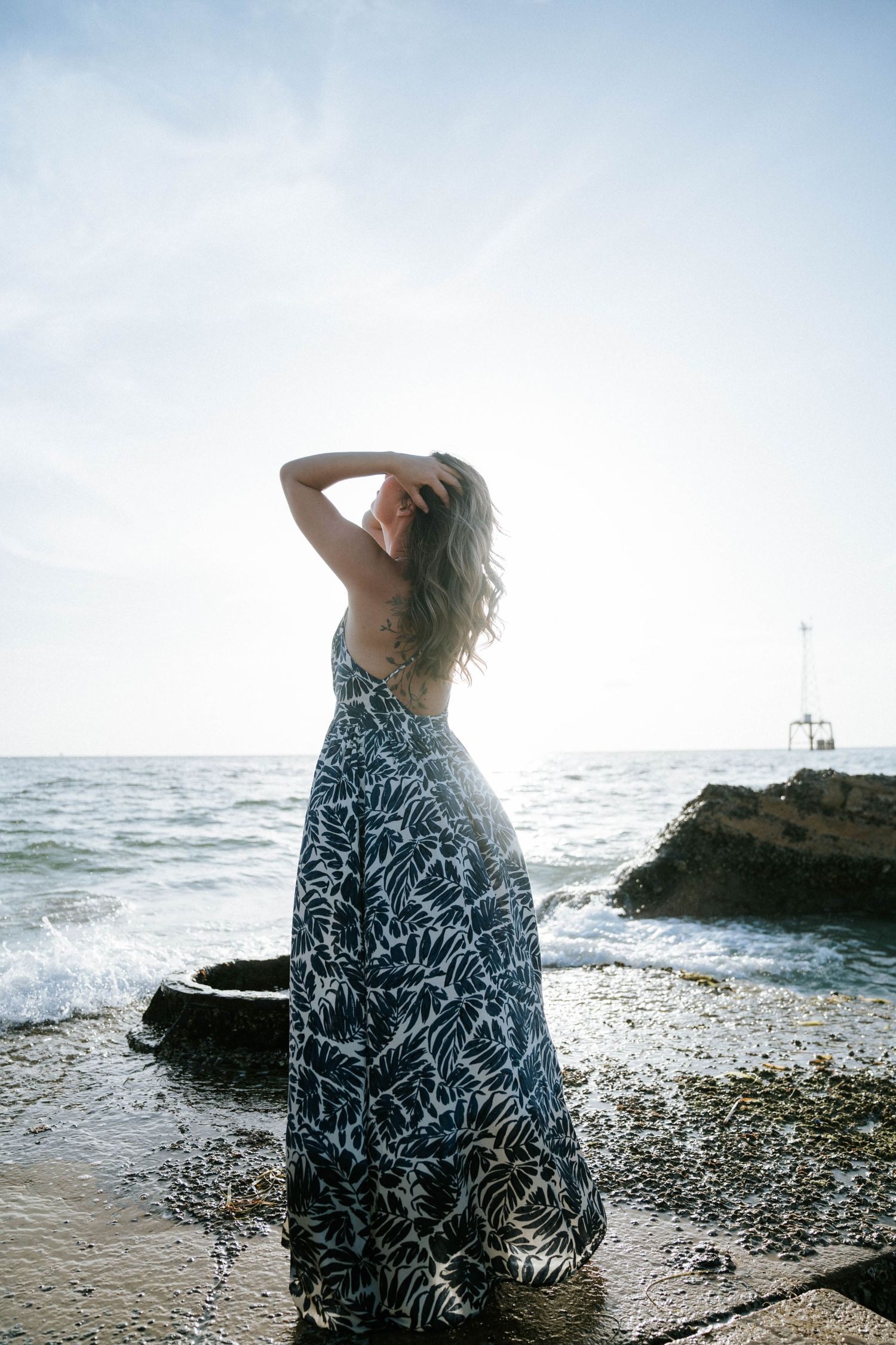 A person in a flowing patterned maxi dress poses on rocky beach at sunset with waves crashing in background.