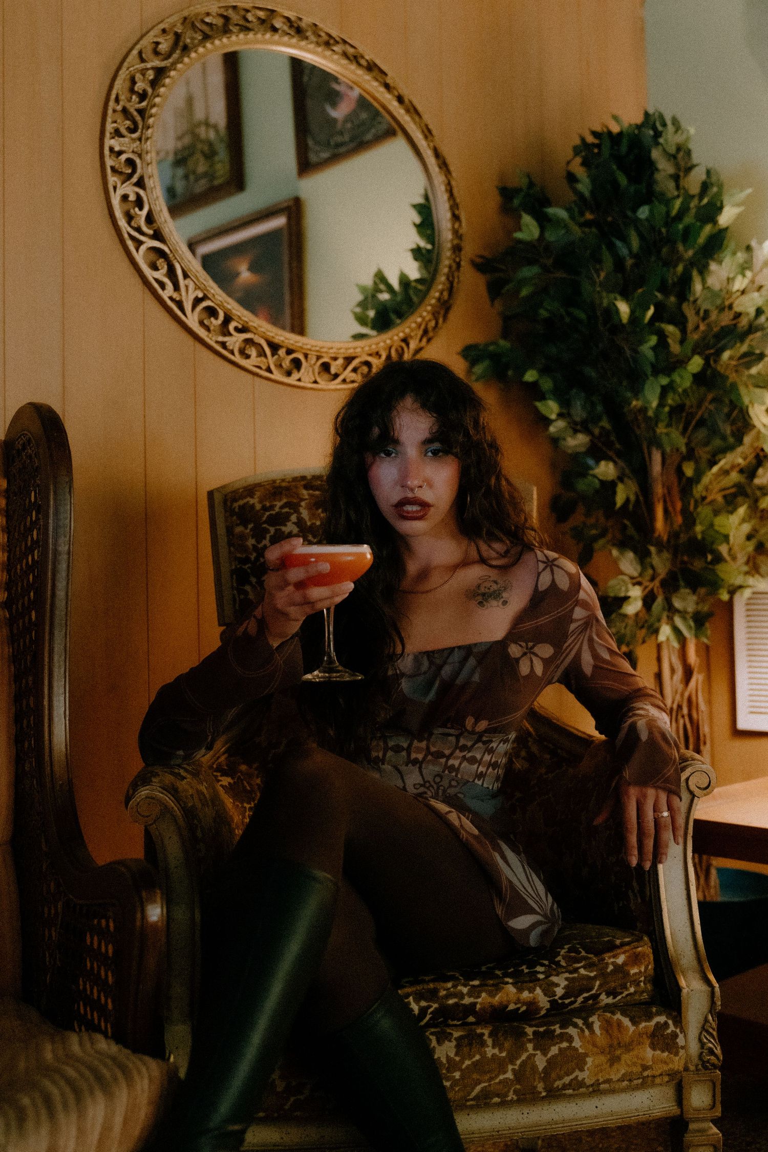 A vintage-style scene shows someone in dark attire holding a cocktail while seated in an ornate chair near a decorative mirror.