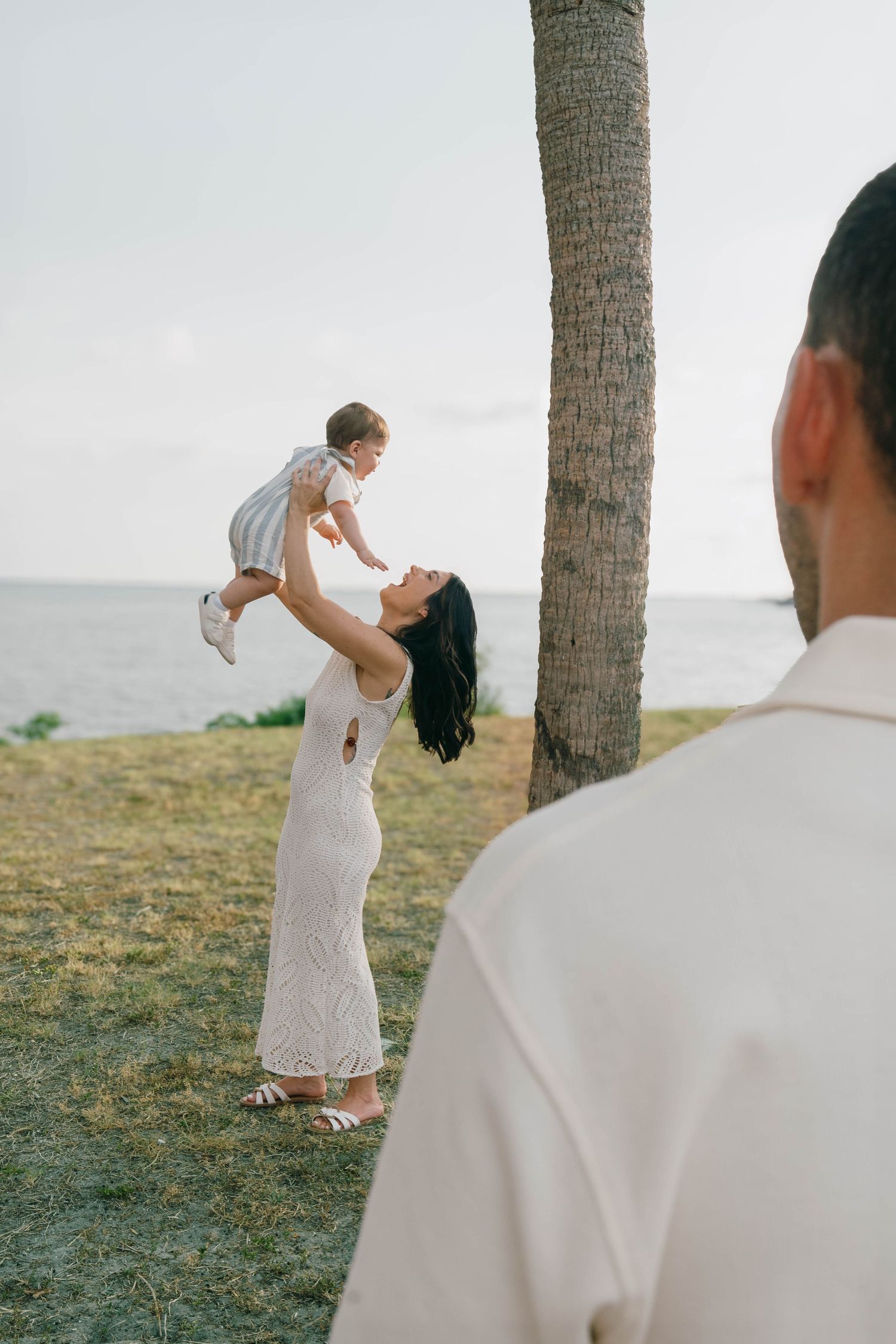 A woman in a white dress lifts a baby near a palm tree by the ocean while a man watches in the foreground.