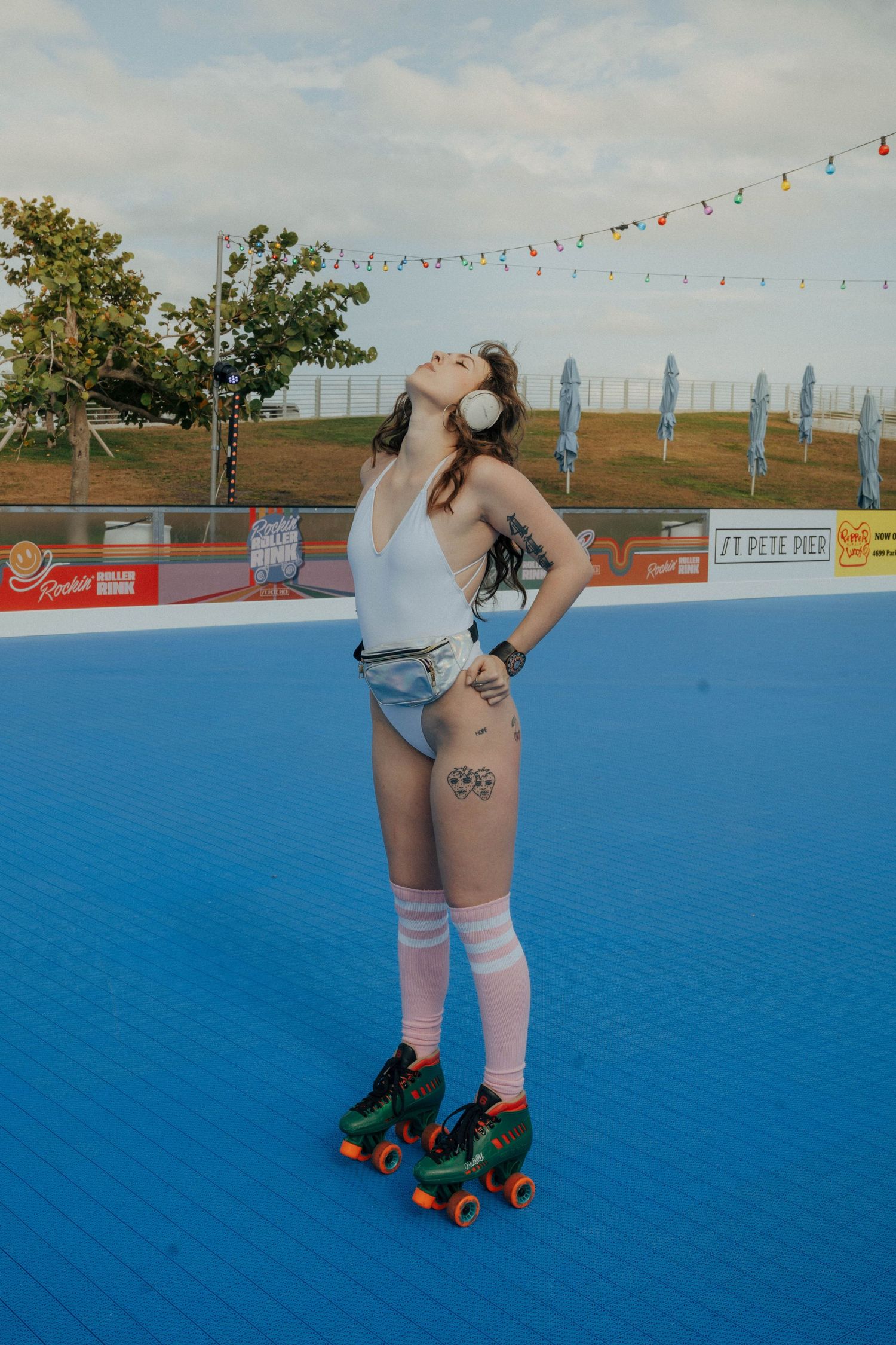 Roller skater in retro shorts and knee socks poses on bright blue outdoor rink with string lights and city skyline.