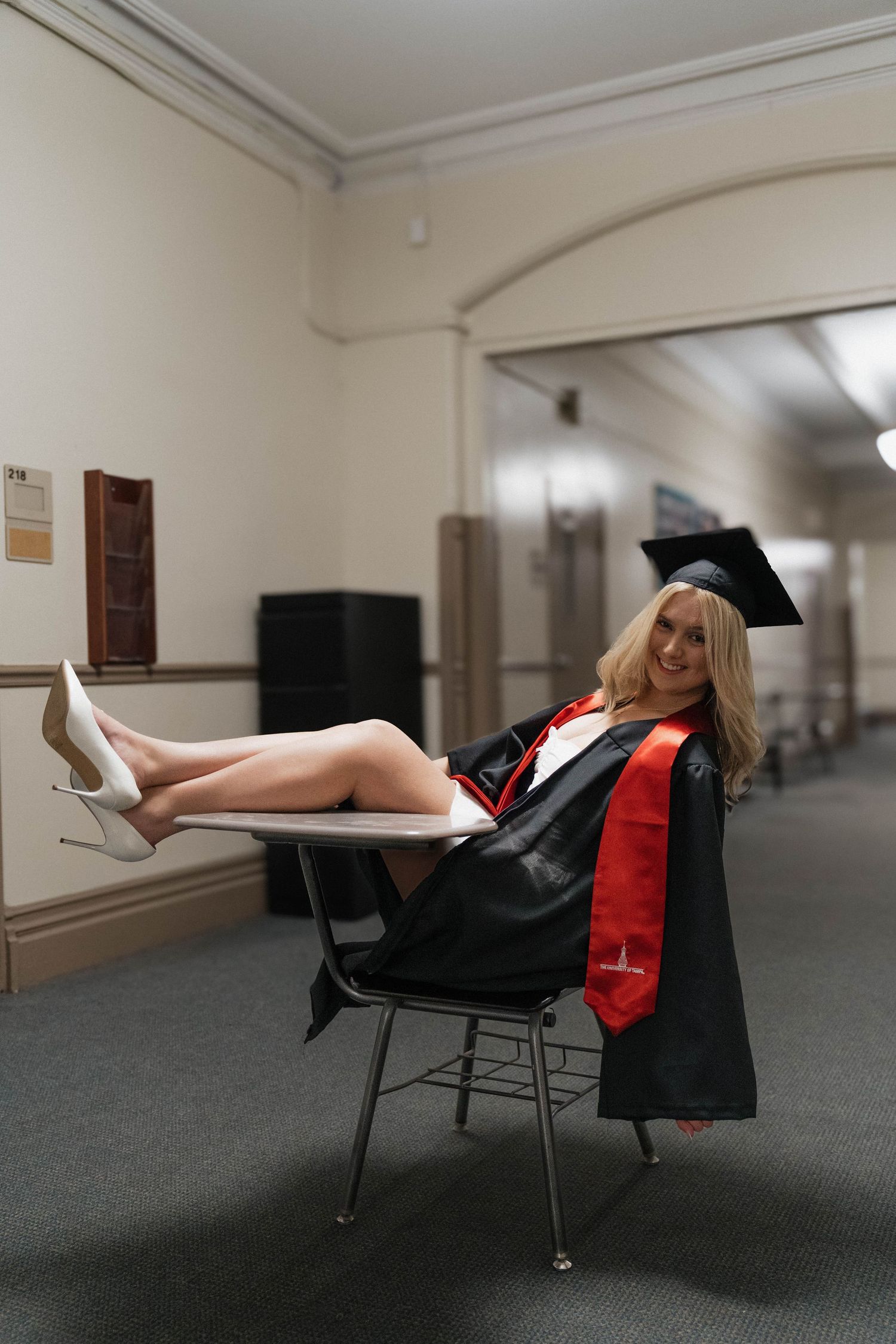 Woman in graduation cap and gown with red stole sits relaxed in chair in university hallway wearing heels.