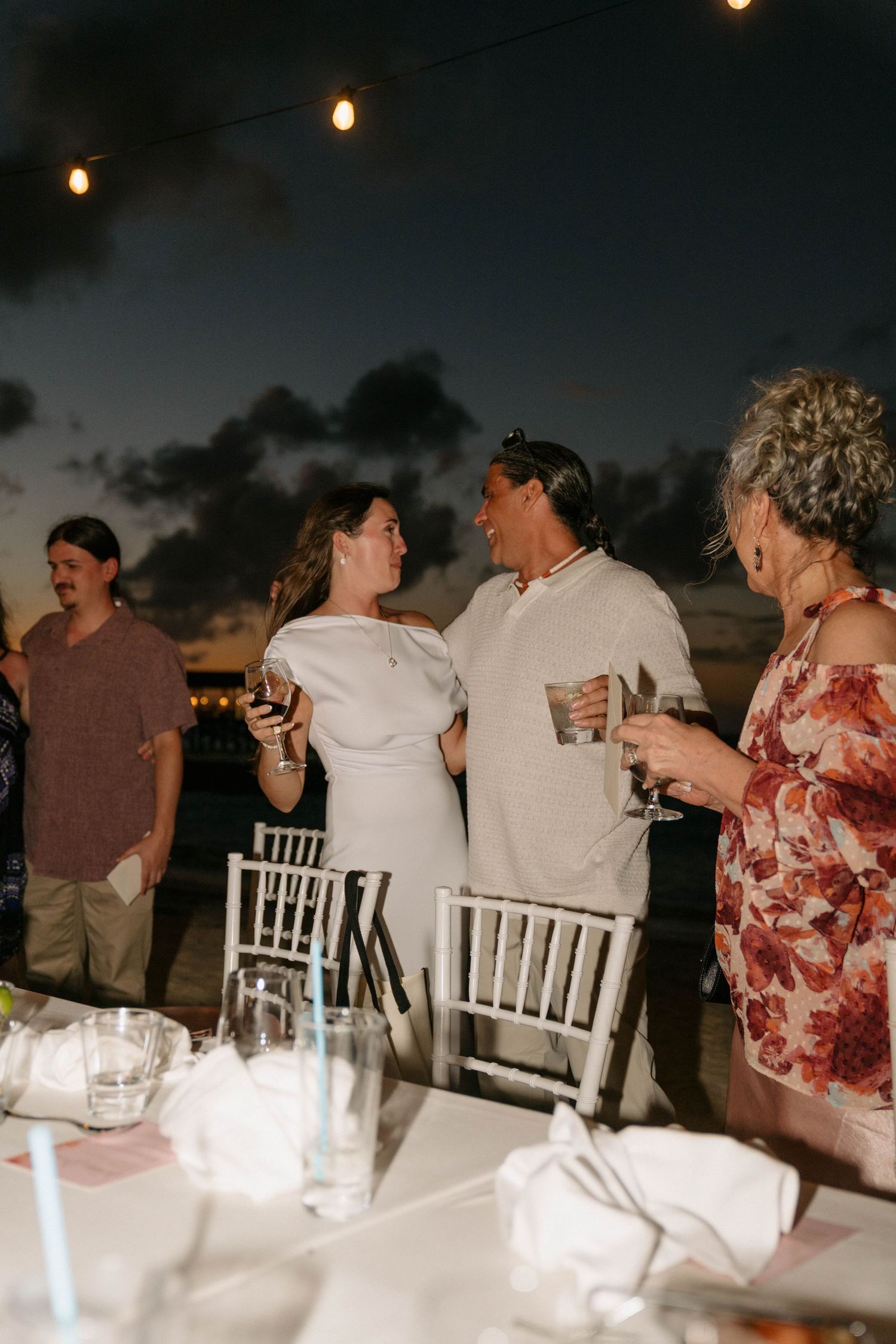 Couple in white attire sharing a romantic moment at an outdoor evening wedding reception with string lights.