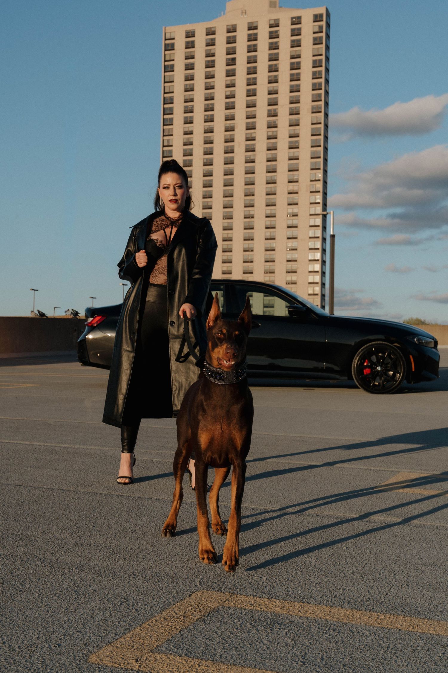 Woman in black leather coat walks Doberman in parking lot with sleek black car and tall building in background.