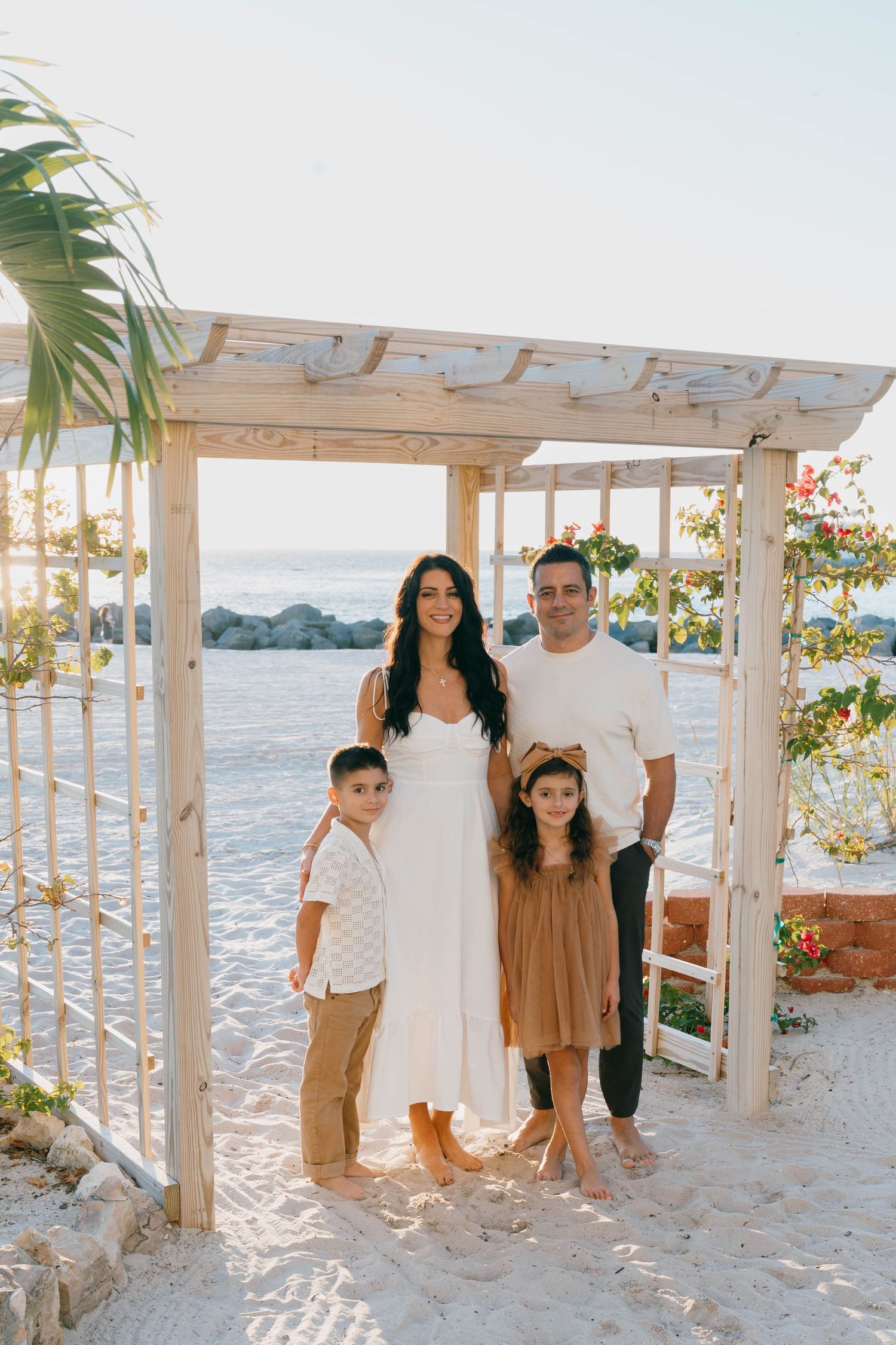Family of four posing under a wooden beach pergola with ocean views, dressed in white and earth tones.