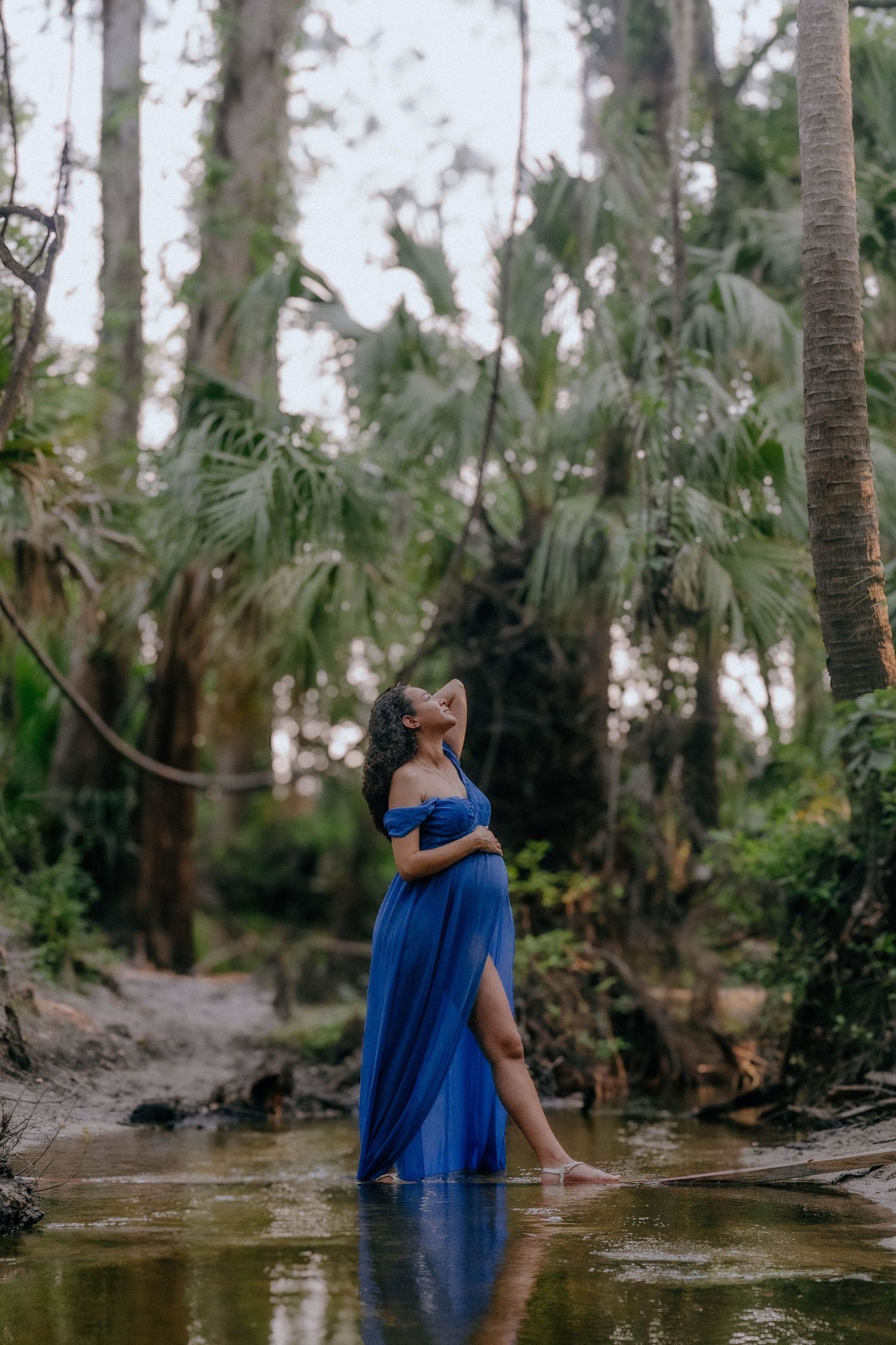 A figure in a flowing blue dress poses in shallow water surrounded by tropical palm trees and forest vegetation.