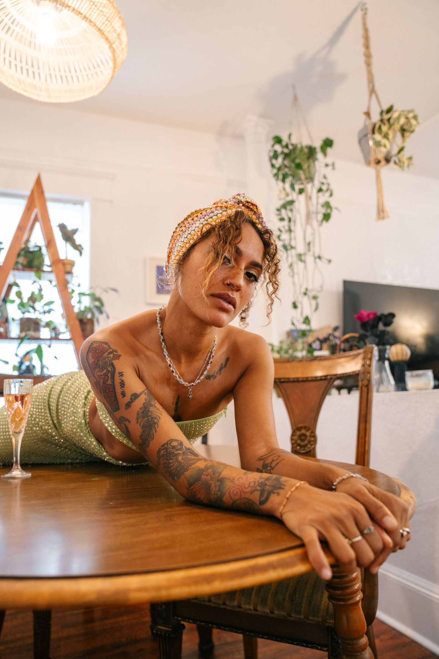Indoor lifestyle portrait showing decorative headscarf, tattoos and green outfit while lounging at wooden table with plants nearby.
