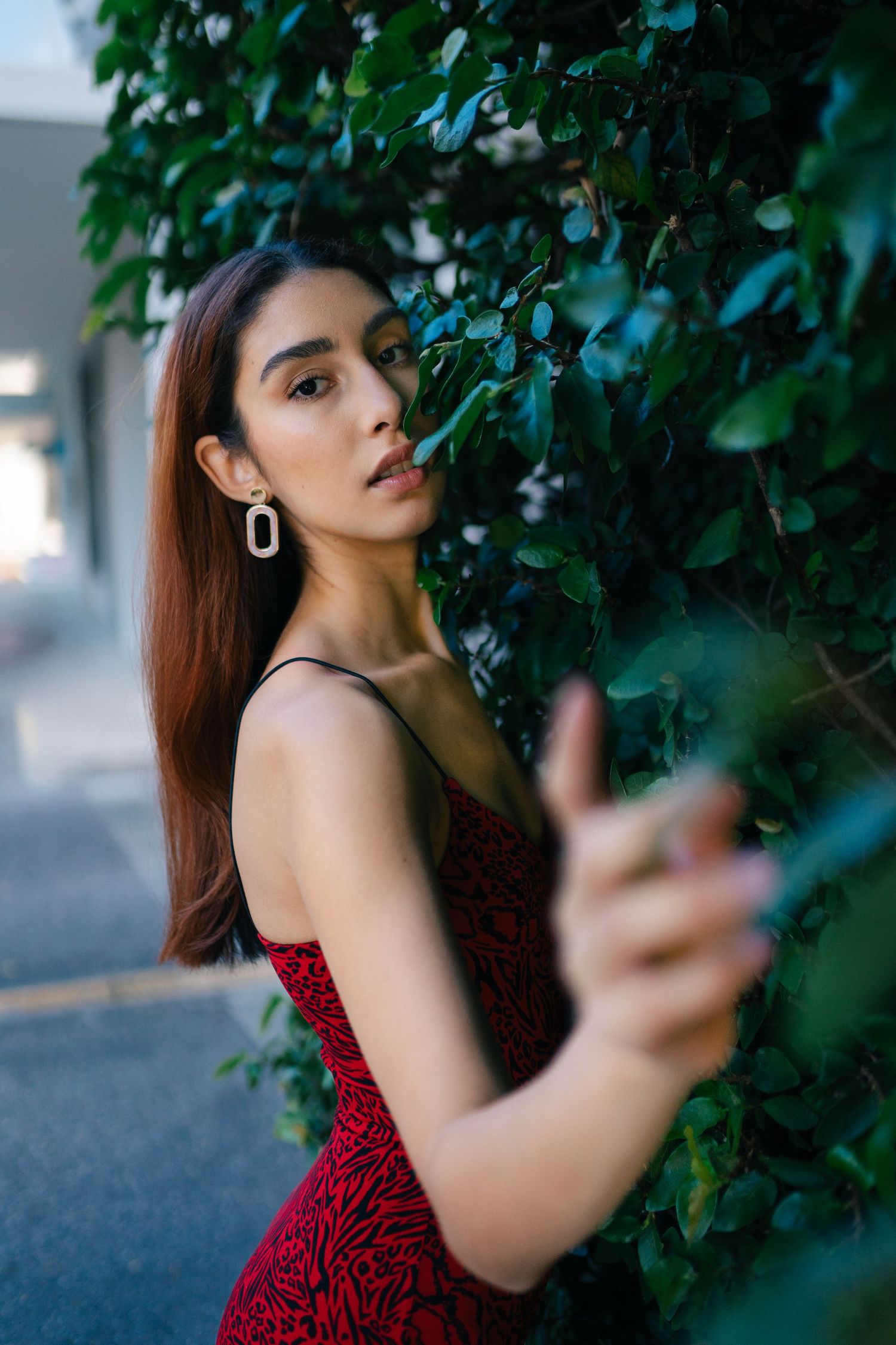 Woman in red dress poses dramatically near green foliage wall.
