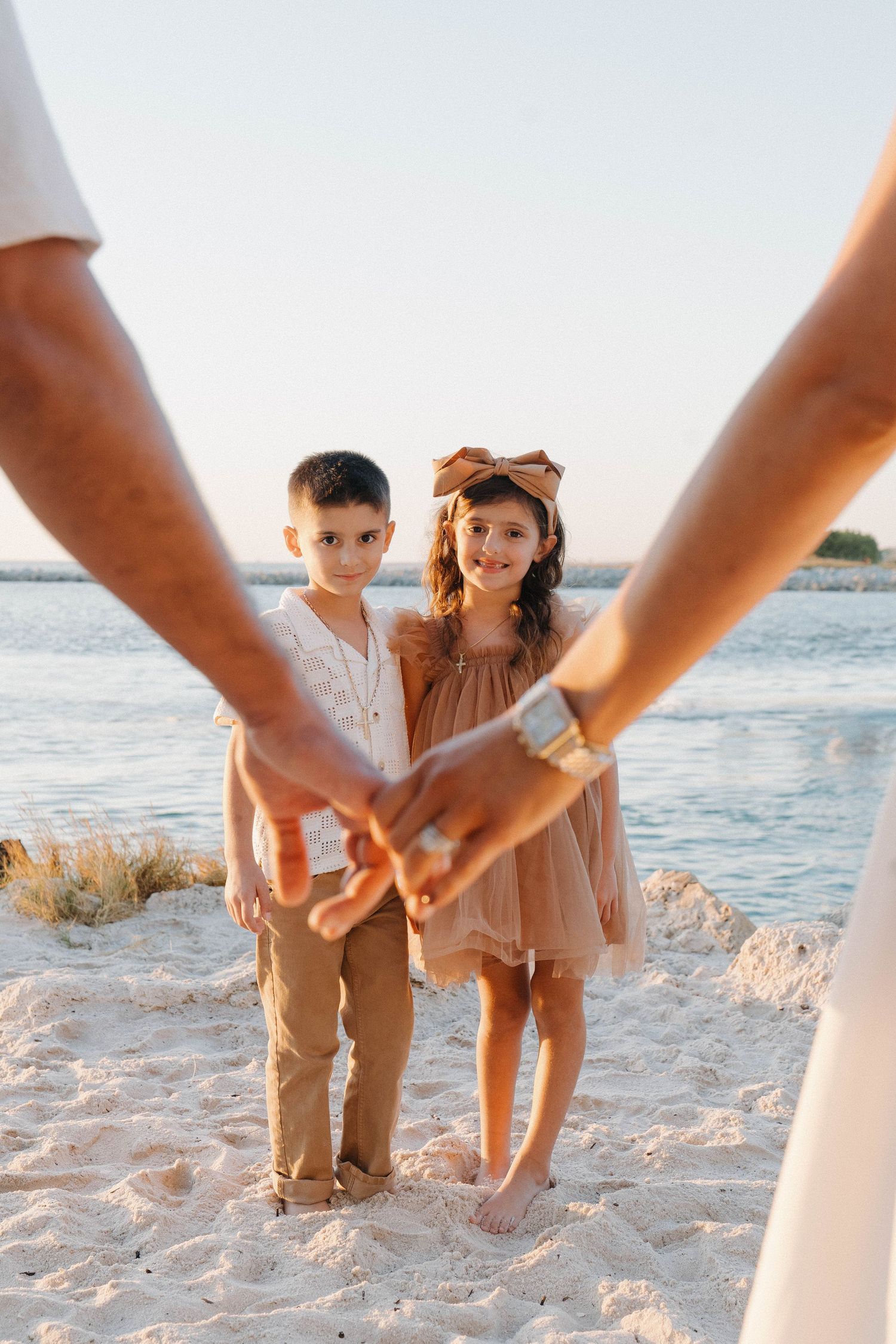 Two children holding hands on a sandy beach at sunset, framed by adults holding hands in the foreground.
