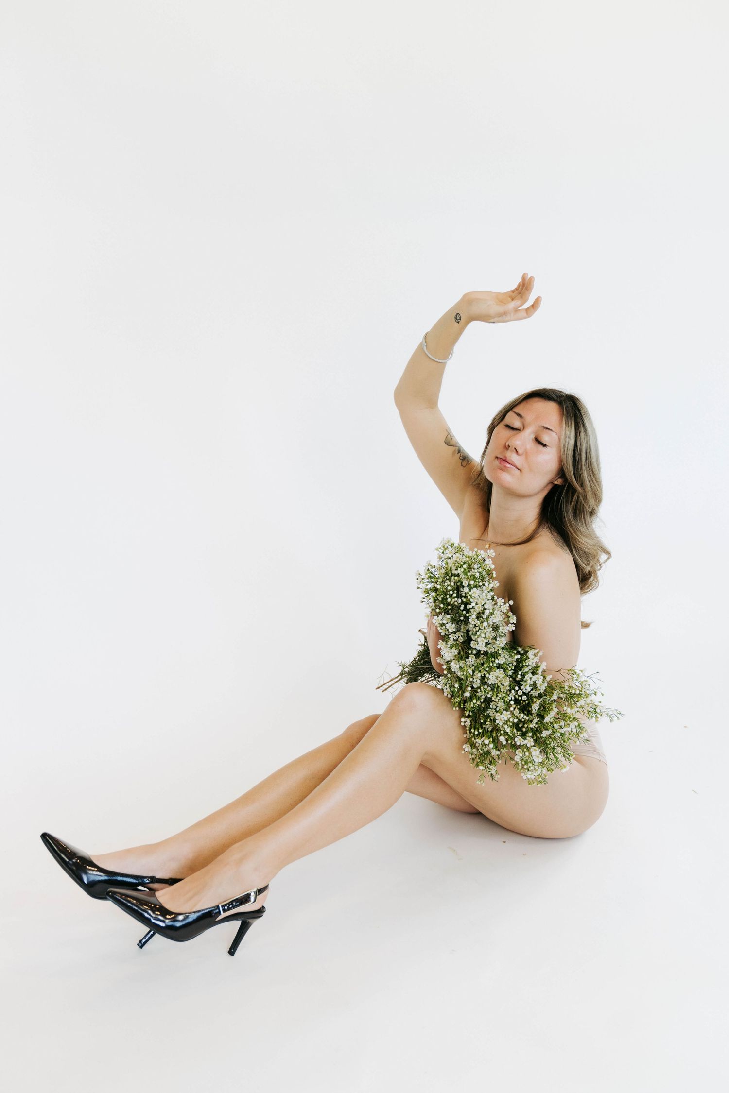 Woman sitting on white background holding white wildflowers, wearing black heels, with one arm raised gracefully.