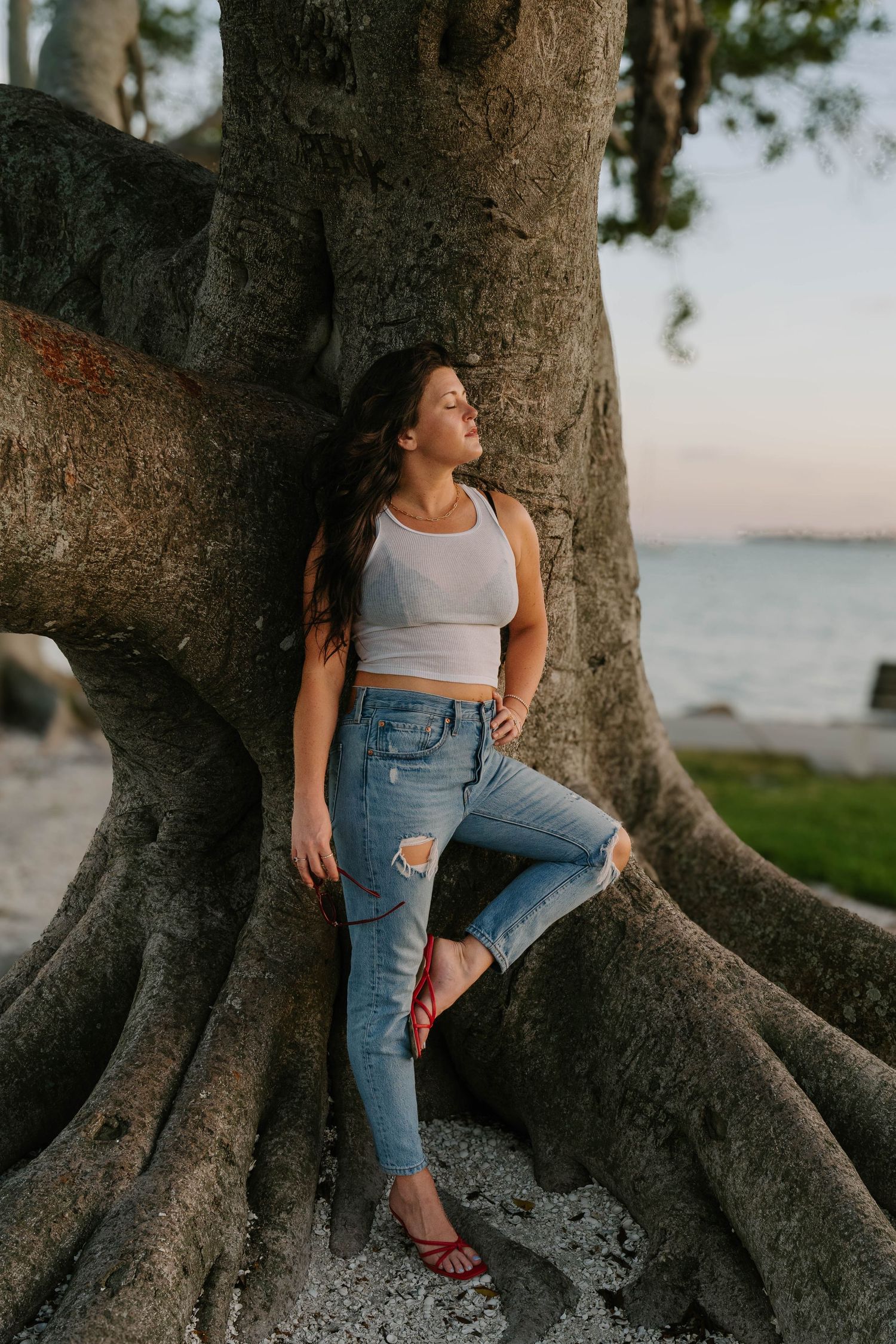Young woman in white crop top and ripped jeans leaning against large tree roots near beach at sunset.