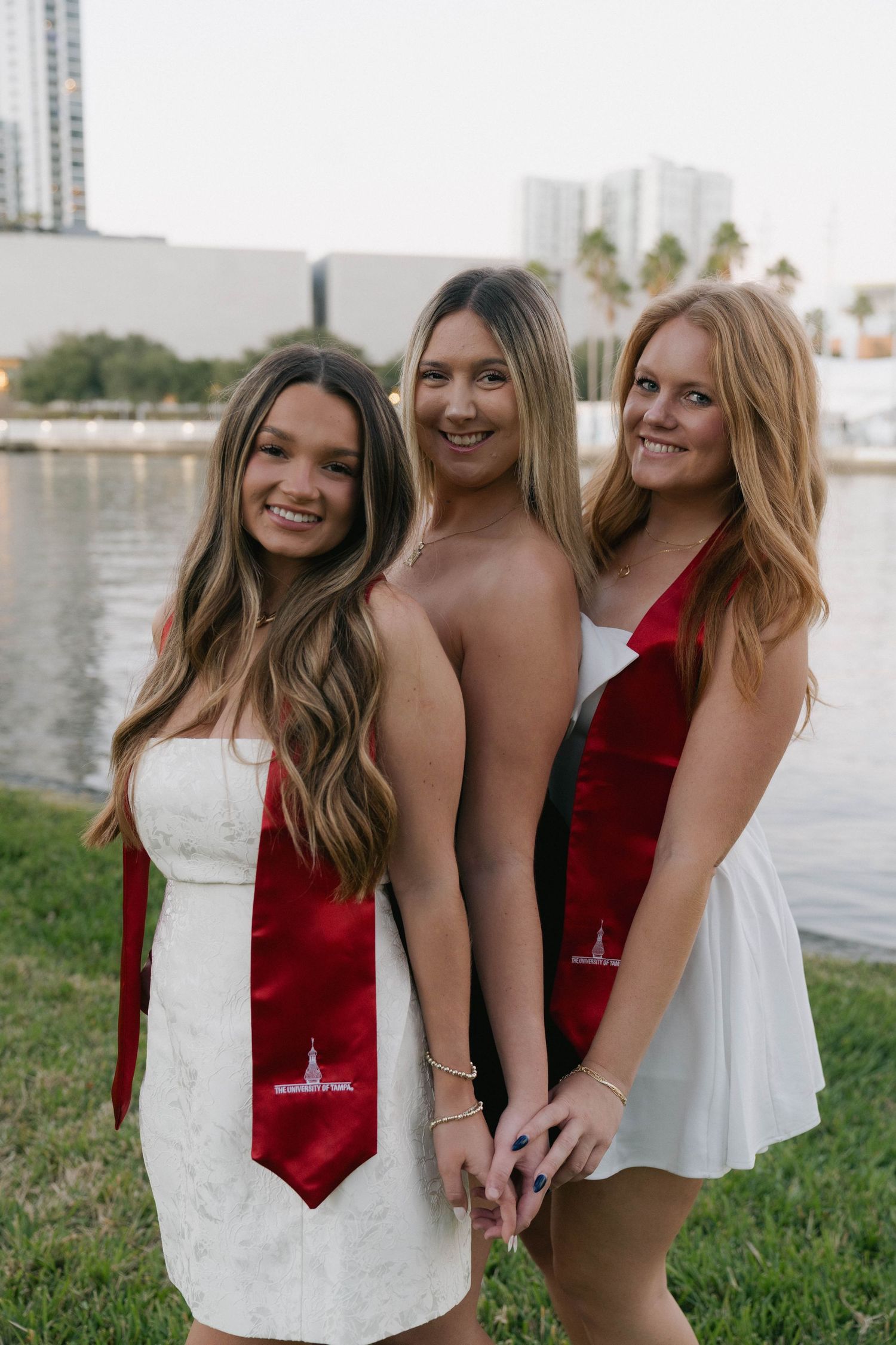 Three smiling young women in white dresses wearing red university graduation stoles pose outdoors near a waterfront.