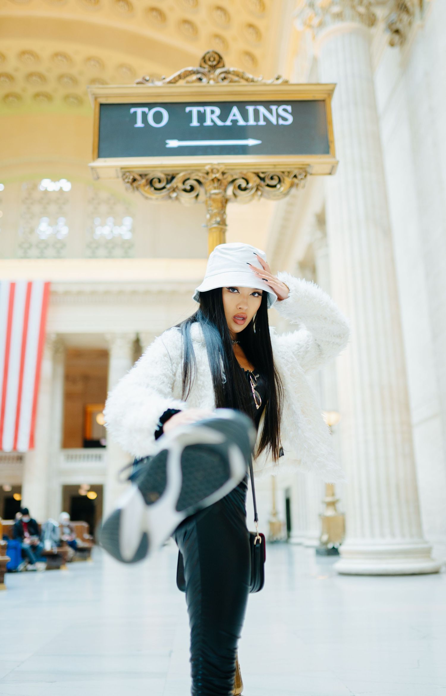 A person in a white hat and fluffy jacket stands beneath a 'TO TRAINS' directional sign in a grand interior space.