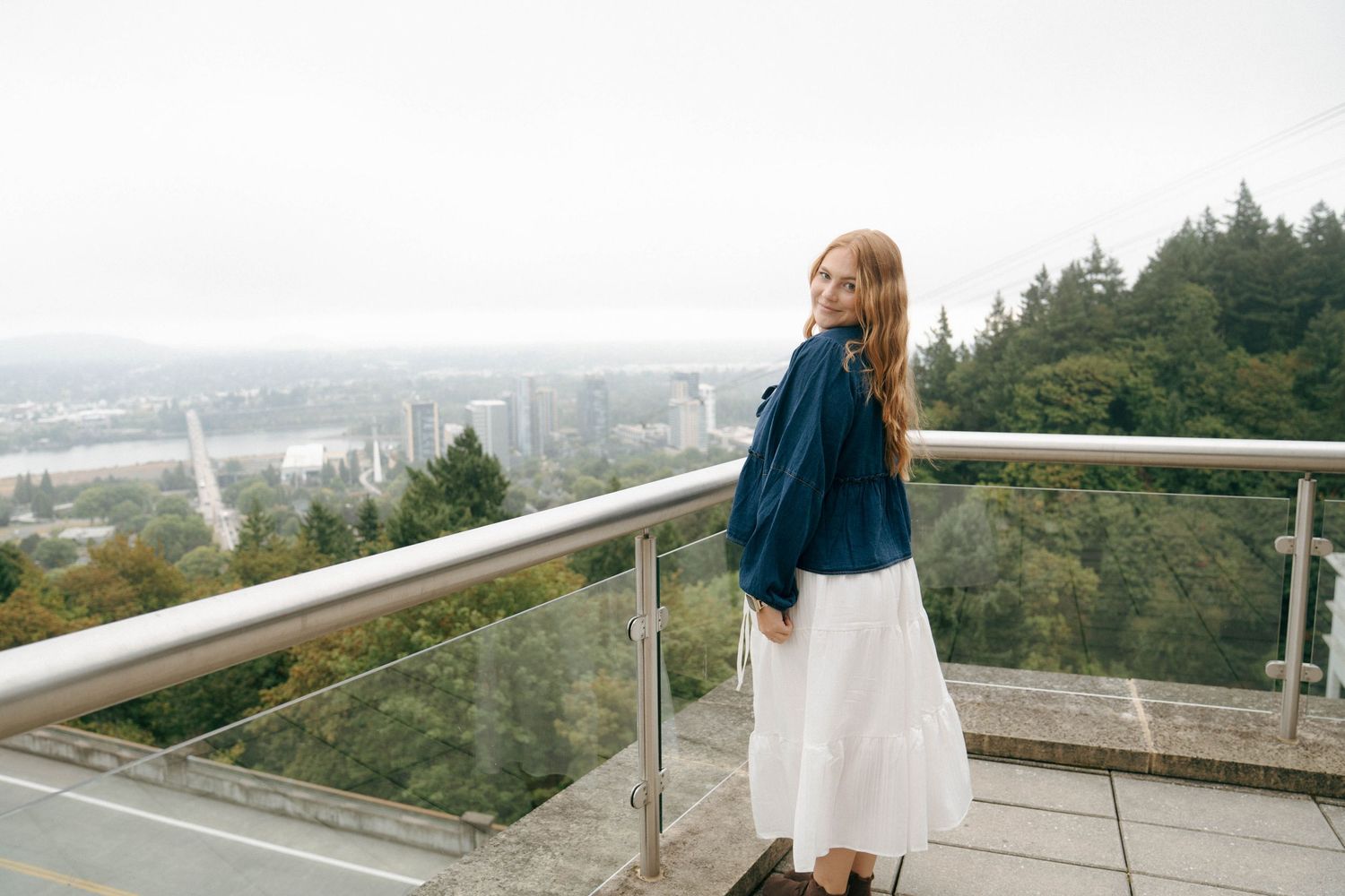 A person in a white dress stands at a scenic overlook with a view of a city and forest below.