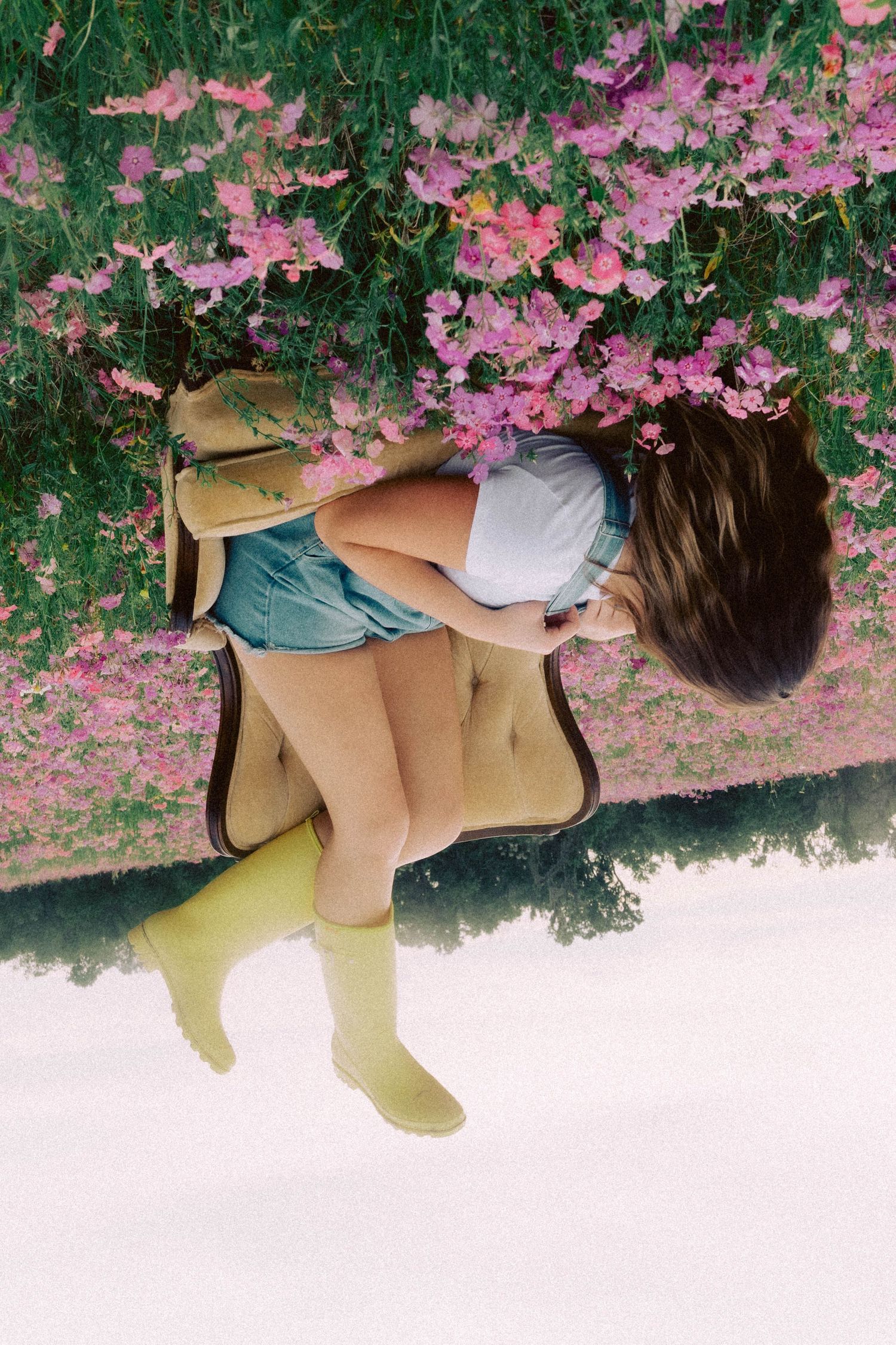 Young girl in yellow boots and blue skirt bending over pink flowering bushes in a dreamy outdoor setting.
