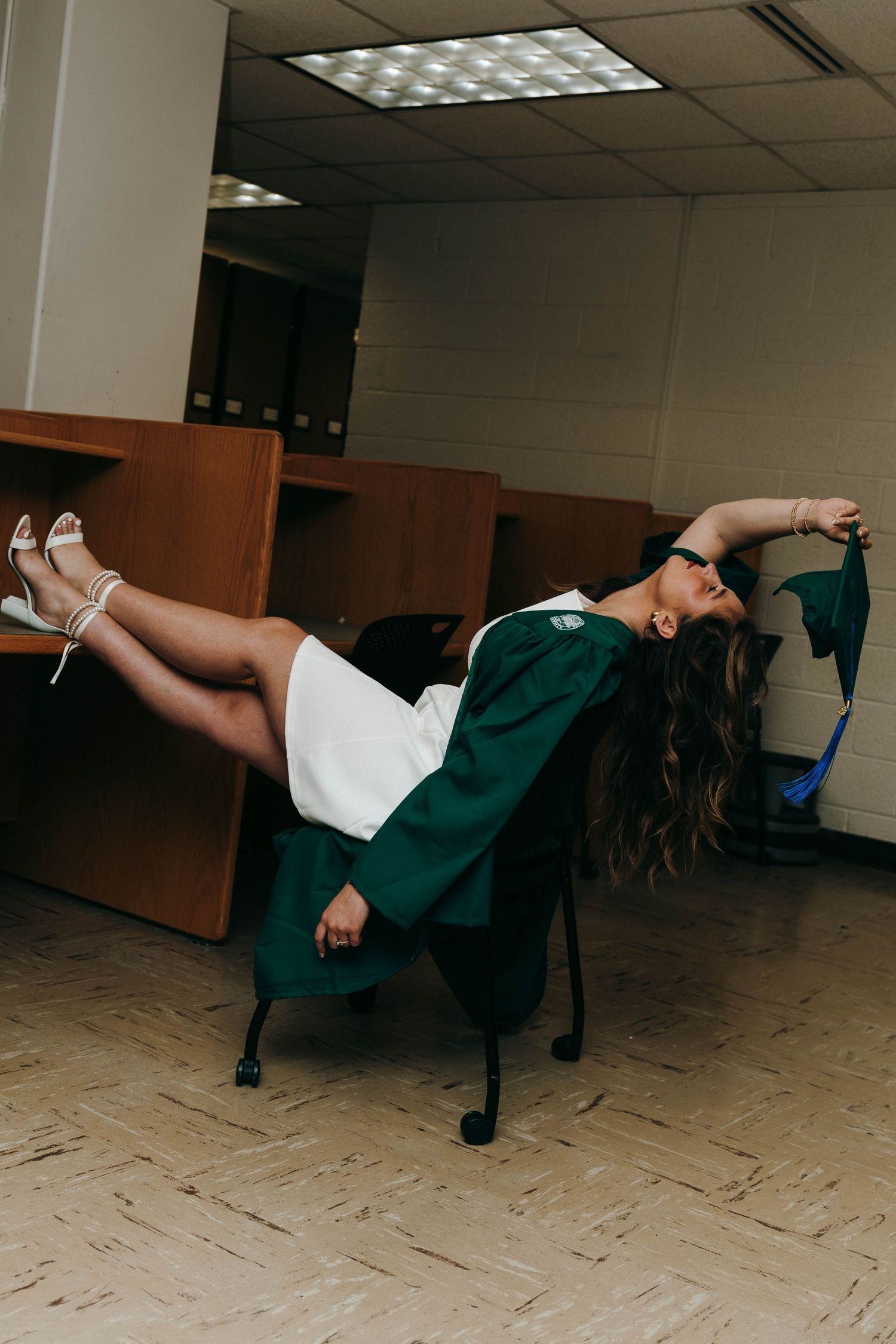 Graduate in green cap and gown dramatically posing in office chair, holding mortarboard, wearing white dress and heels.