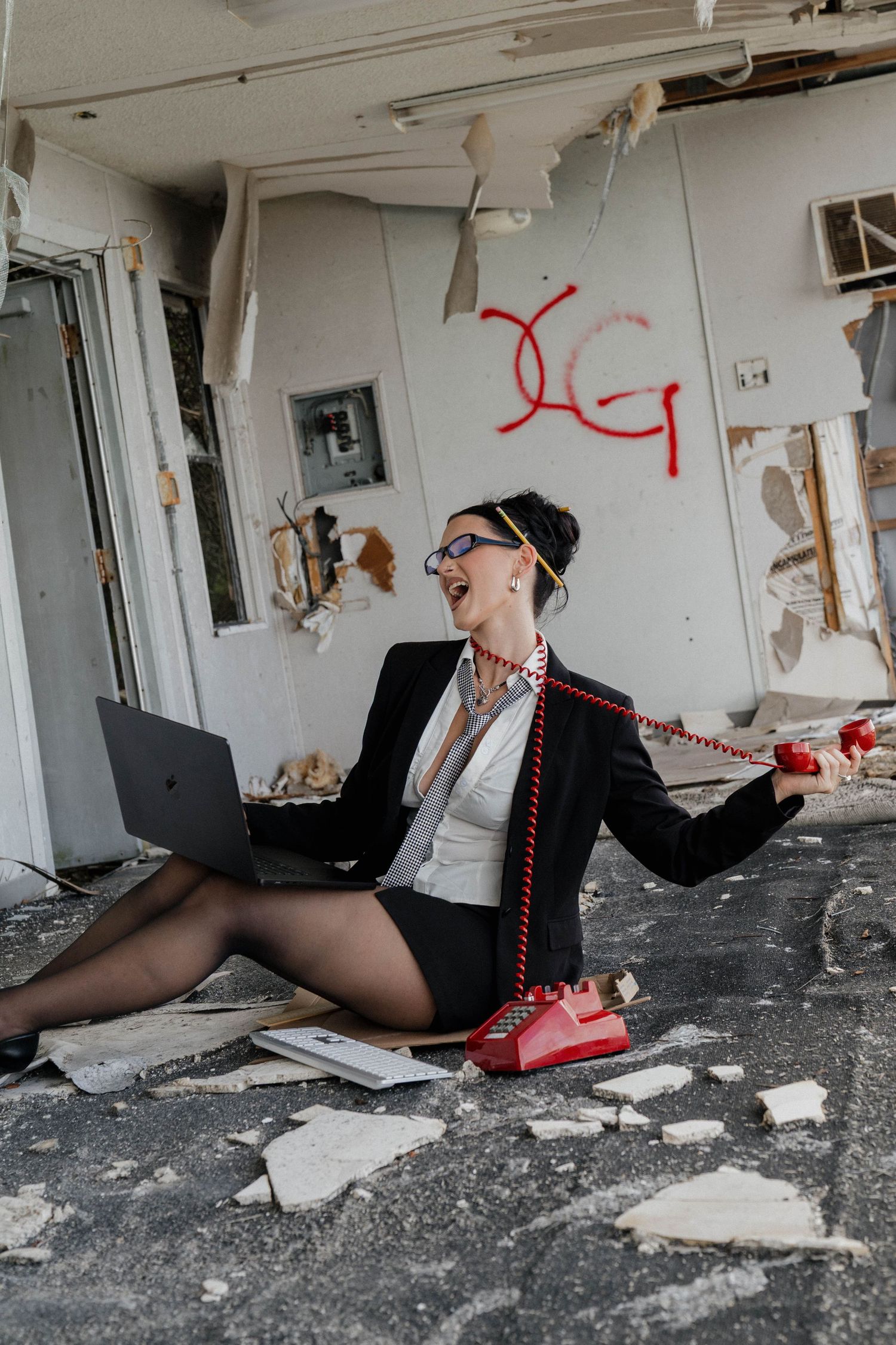 Woman in business attire sits on debris-covered floor with laptop and red phone in abandoned building.