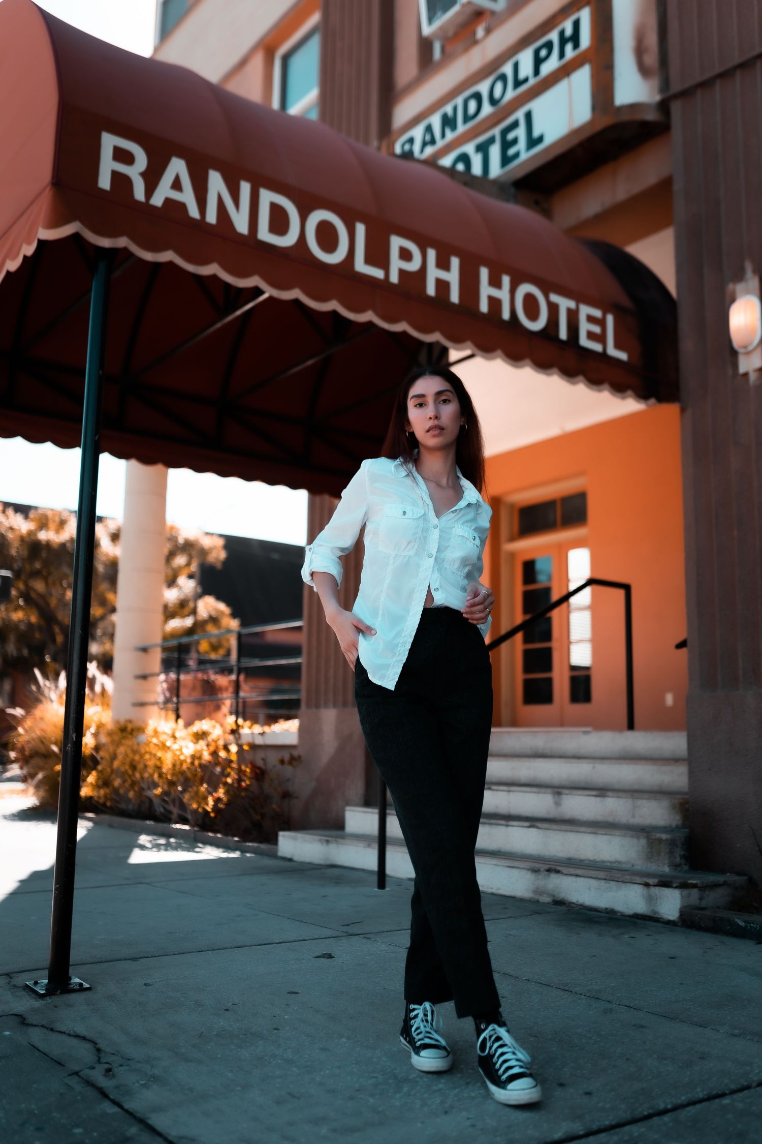 A stylishly dressed person poses in front of the Randolph Hotel entrance wearing a white shirt and black pants with sneakers.