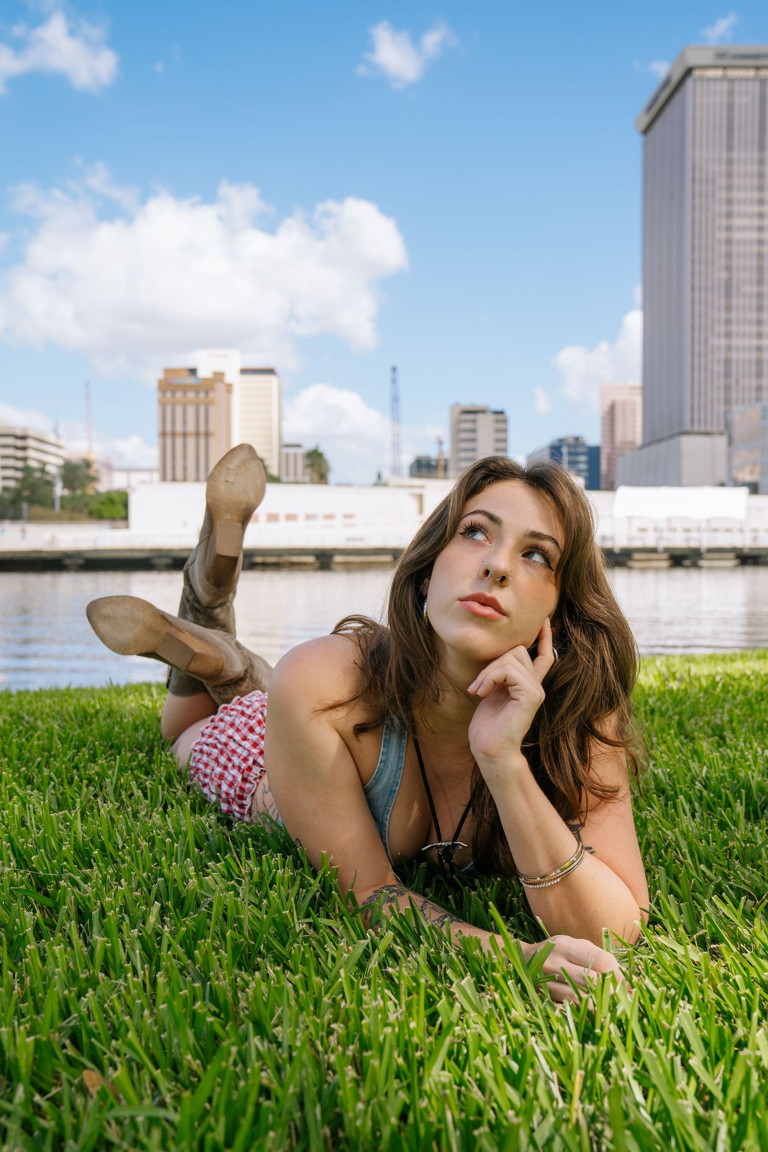 Woman relaxing on green grass by riverside with city skyline and buildings in background on sunny day.