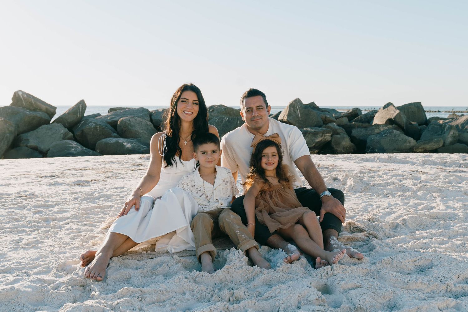 Family of four smiling together on a sandy beach with rocky shoreline backdrop on a sunny day.