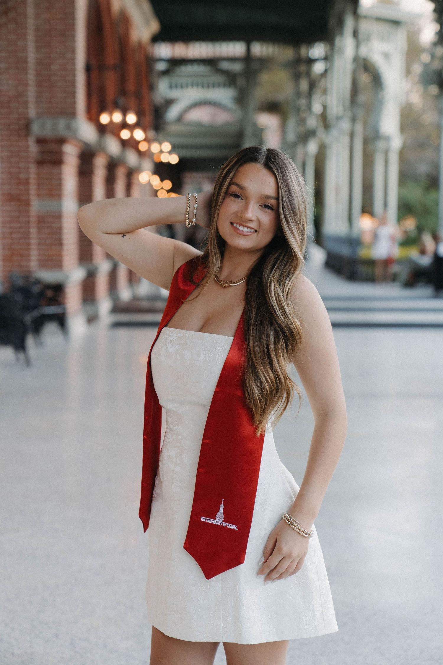 Smiling young woman in white dress wearing red graduation stole poses in elegant brick arcade building.