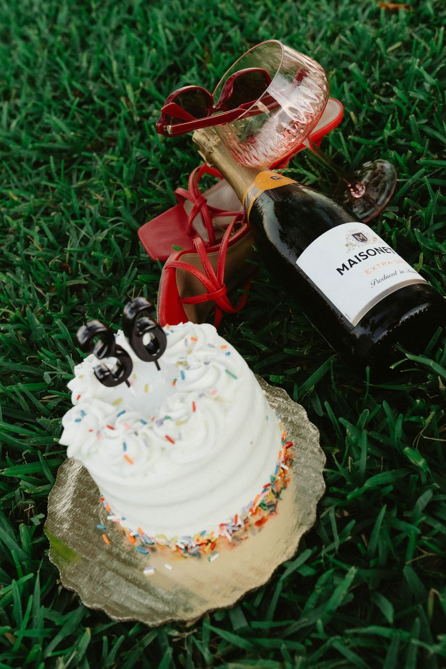 A 30th birthday celebration with a white frosted cake, sprinkles, number candles, and a Maison bottle on green grass.