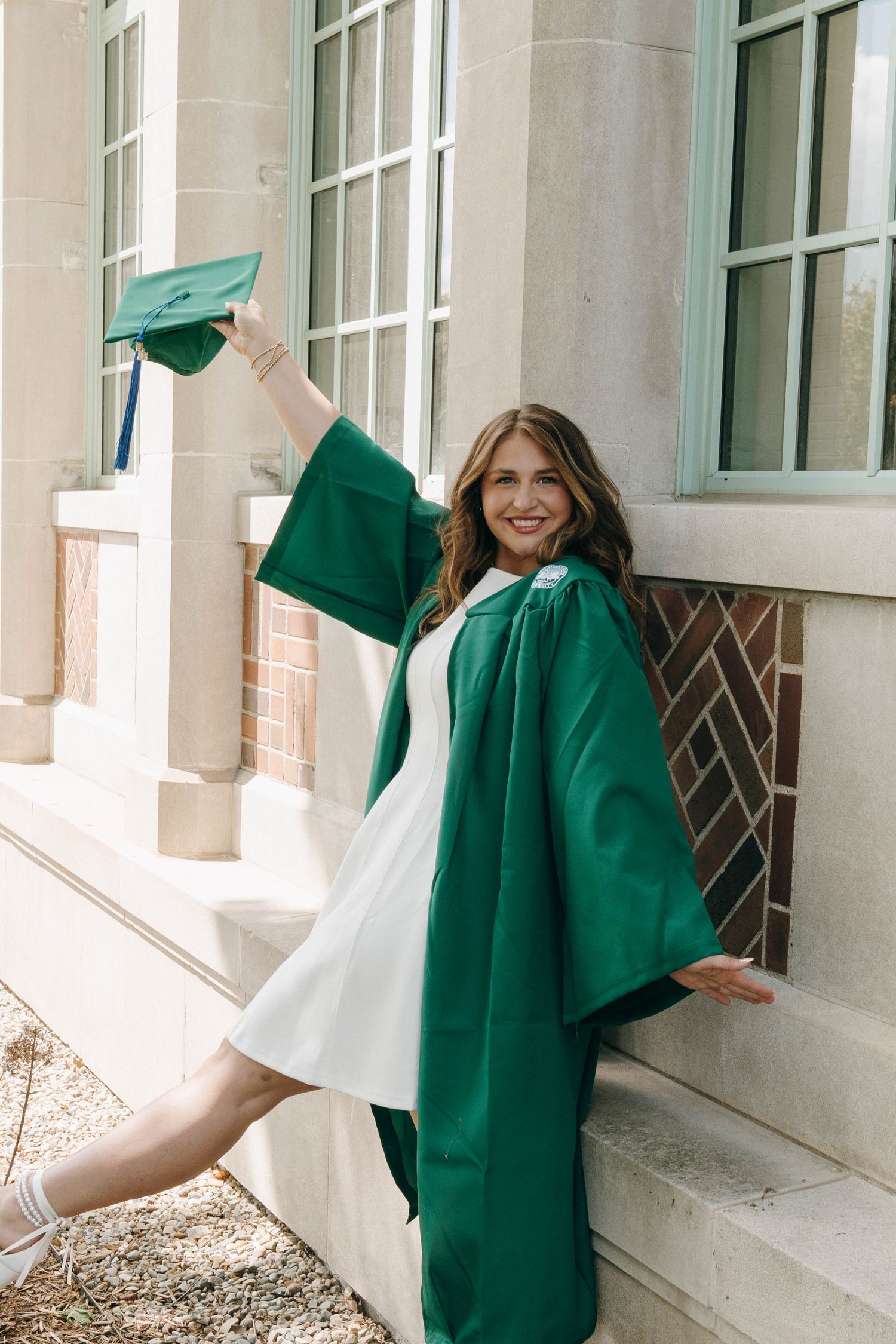 Young woman in green graduation cap and gown celebrating outdoors, raising cap with a joyful smile near a brick building.