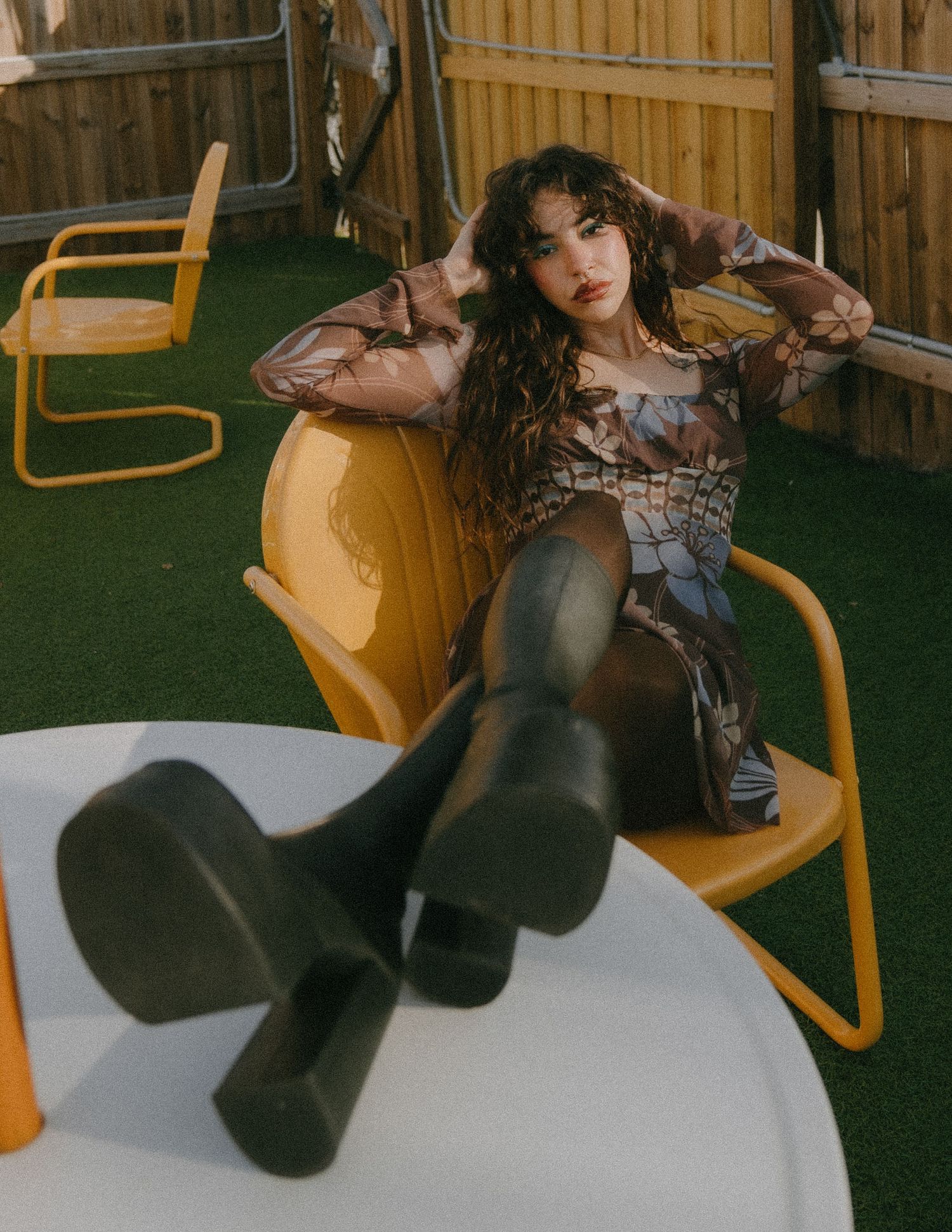 Stylishly dressed figure lounges in retro yellow chairs on artificial turf with wooden fence backdrop.