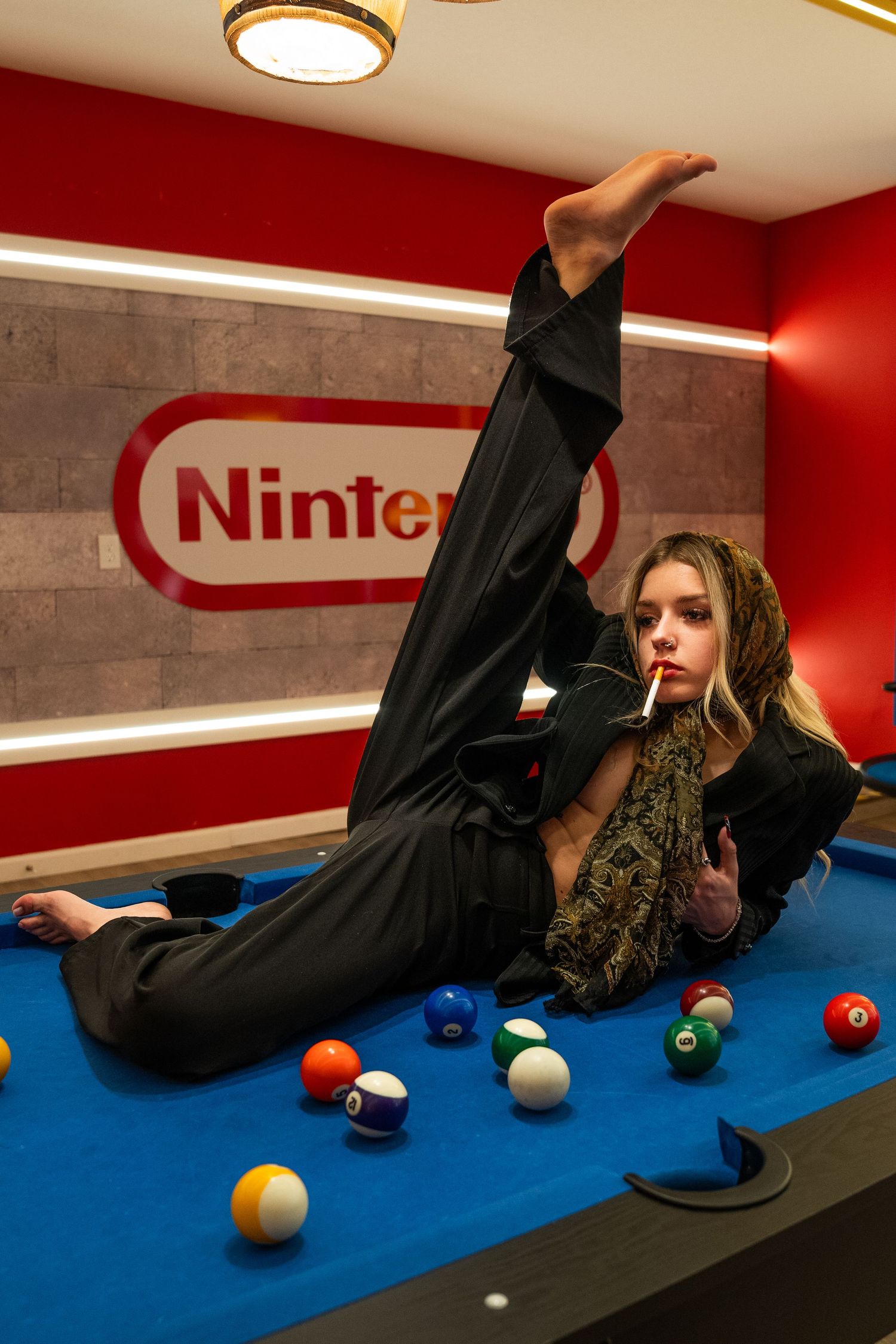 A person in black attire poses on a blue pool table with colorful billiard balls beneath a Nintendo logo on a red wall.