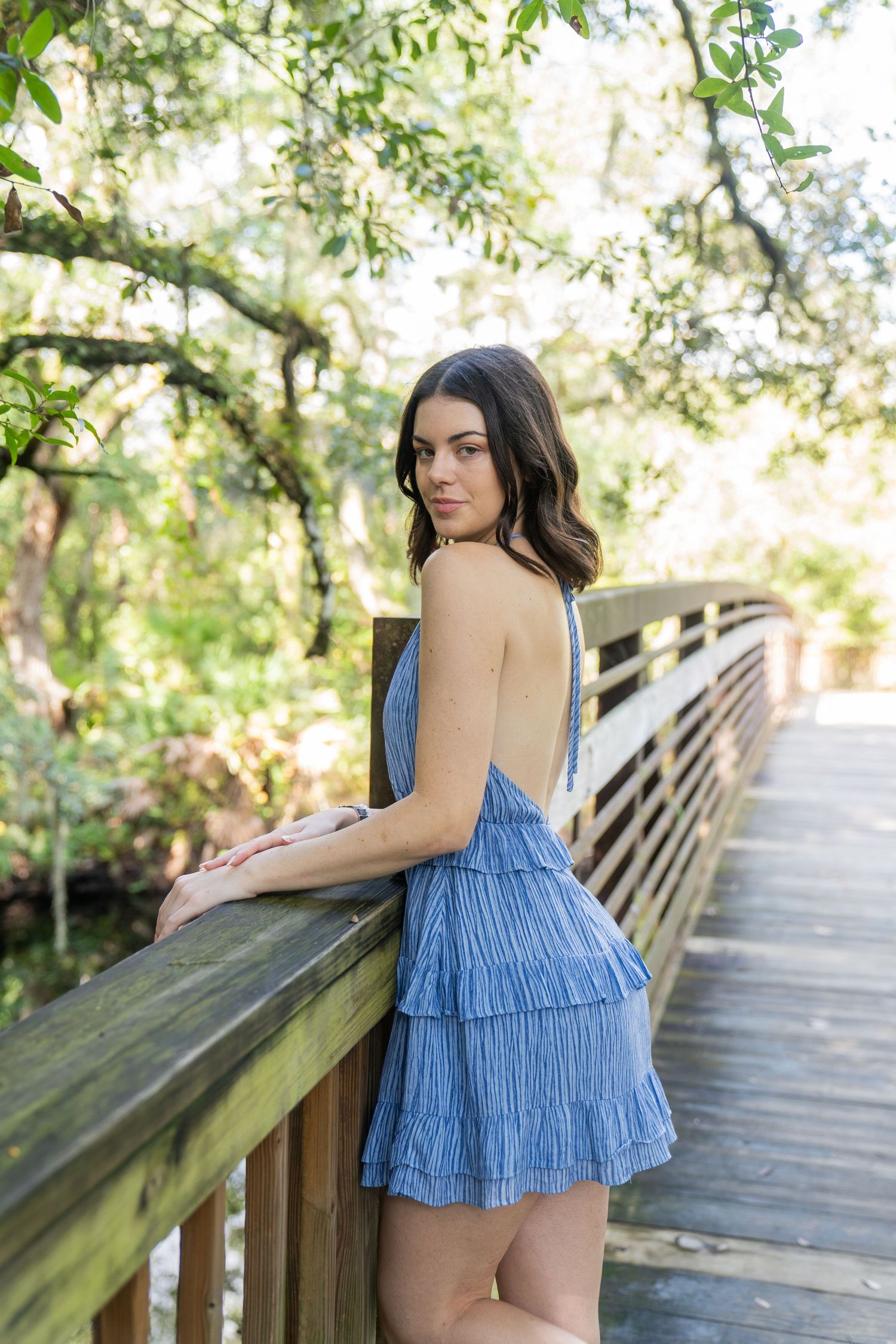 A blue tiered ruffle dress catches sunlight on a wooden bridge walkway surrounded by lush greenery.
