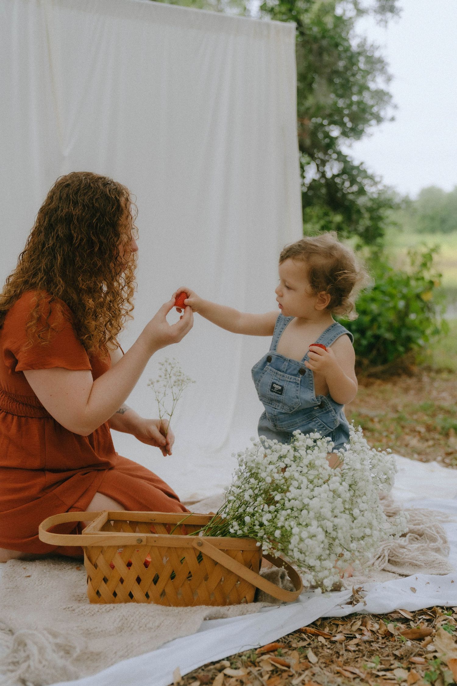 A touching outdoor picnic scene with a child and adult sitting on a white blanket with flowers and a woven basket.