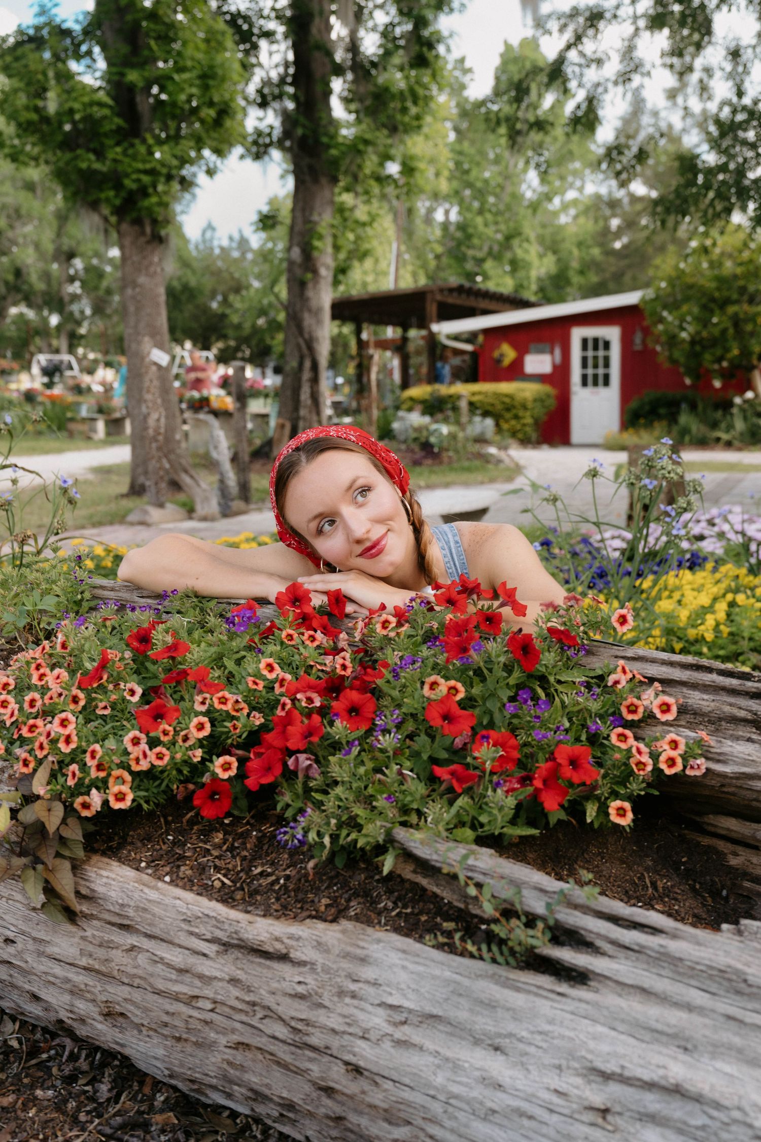 Young woman in red bandana rests chin on hands over colorful petunias in a rustic garden planter.