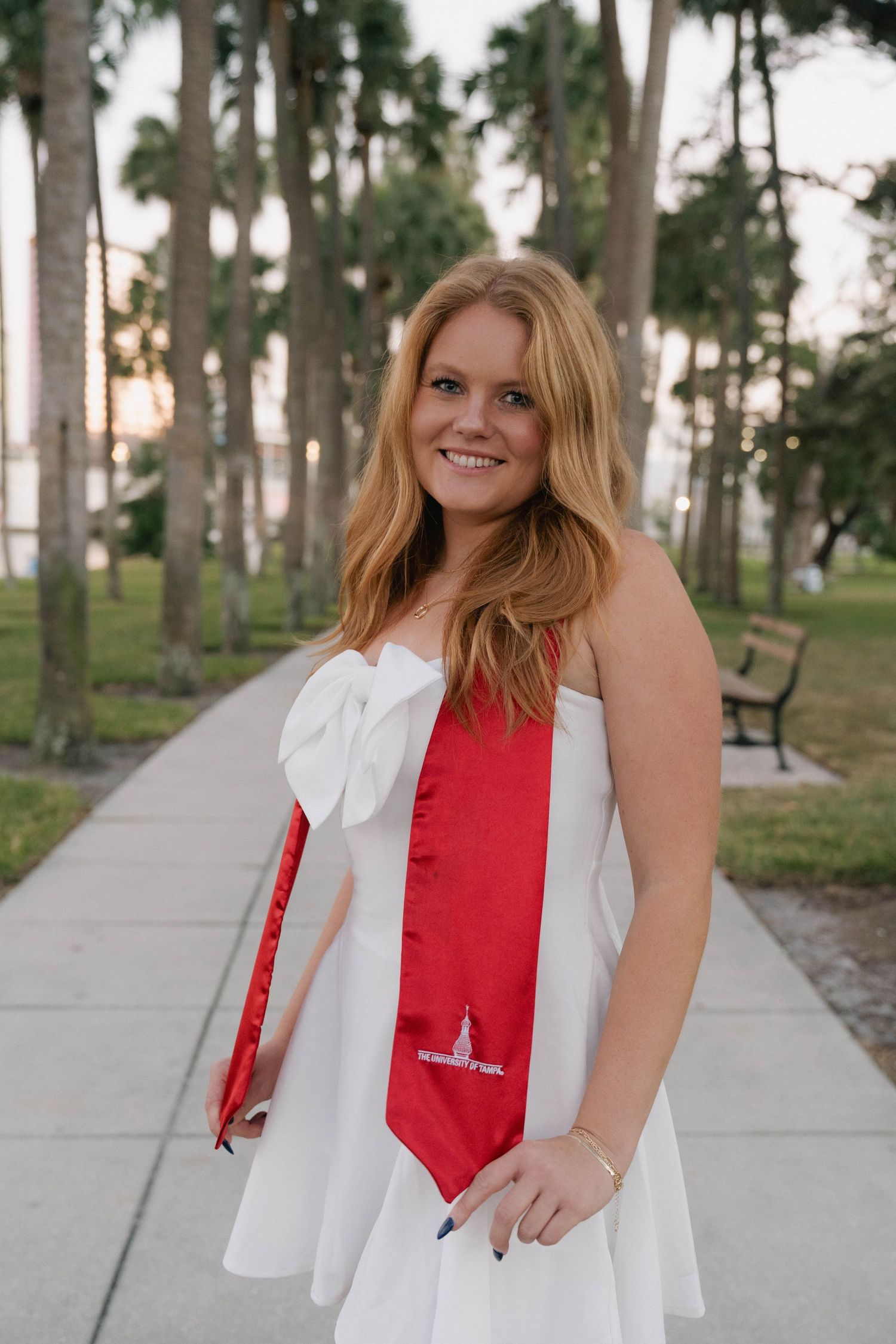 Smiling graduate in white bow dress with red stole posing on palm tree-lined walkway outdoors.