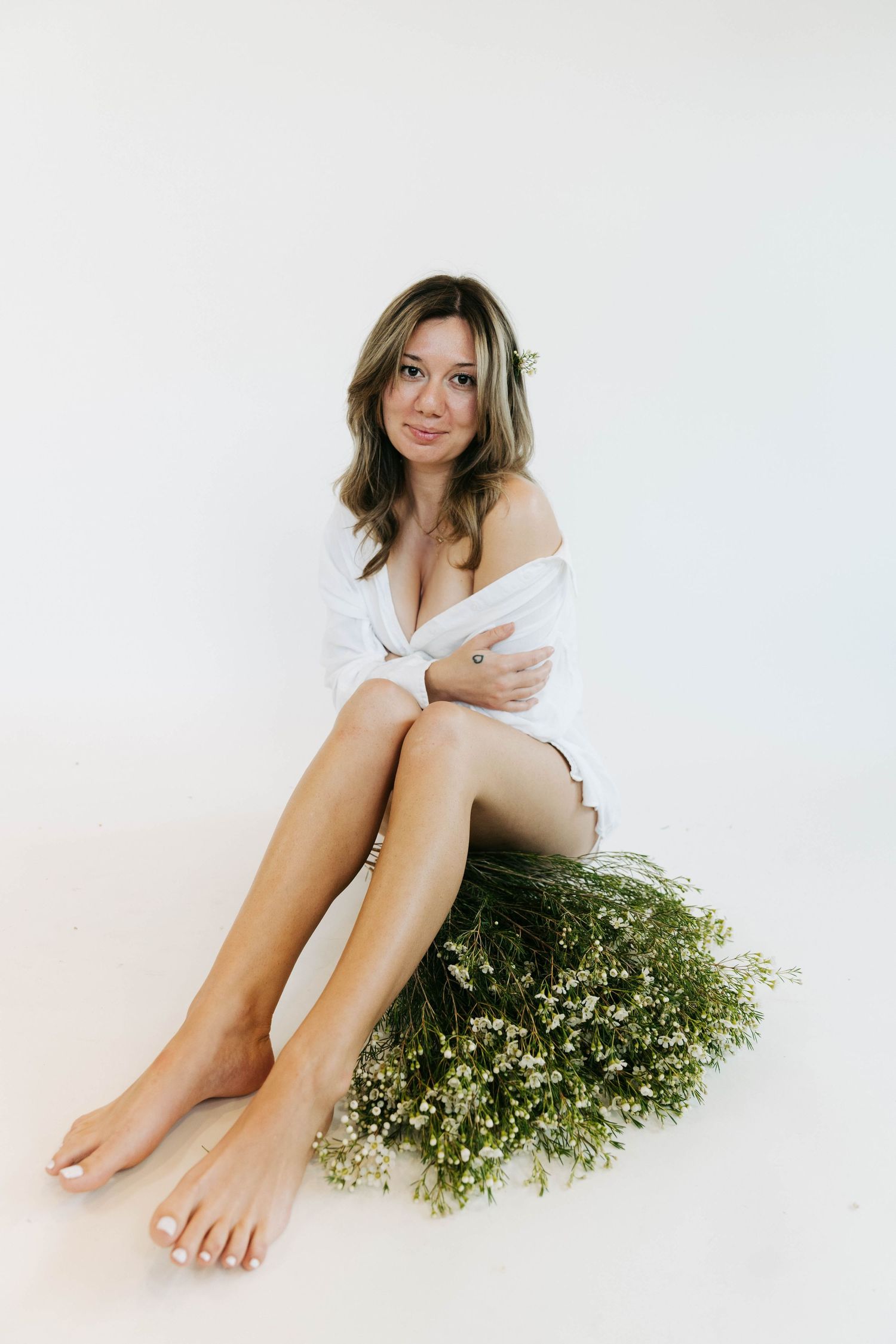 Woman in white shirt sitting barefoot on white floor with bouquet of wildflowers, posing for portrait photo.
