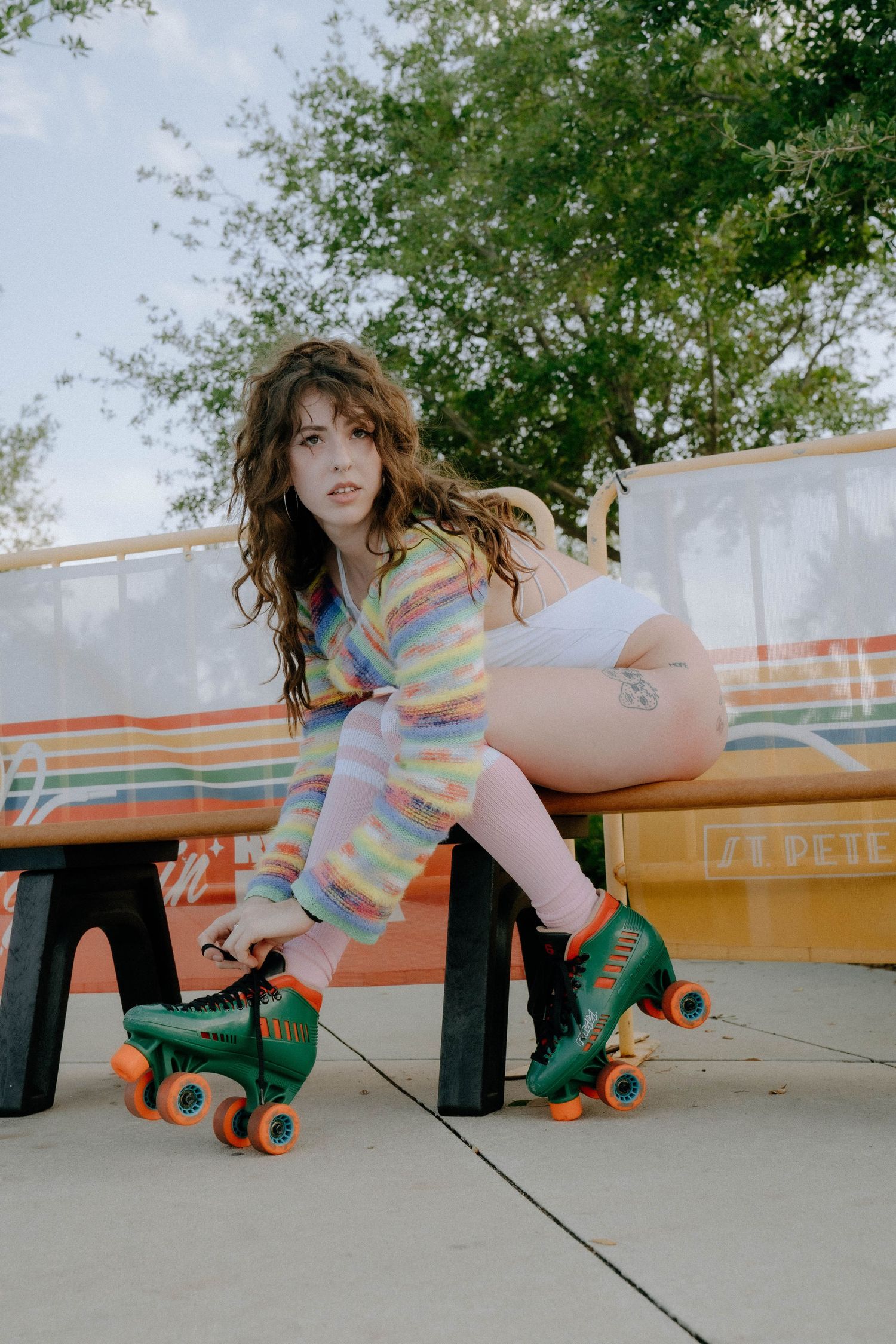 Young woman with curly hair wearing tie-dye outfit and green roller skates sits on bench near colorful retro mural.