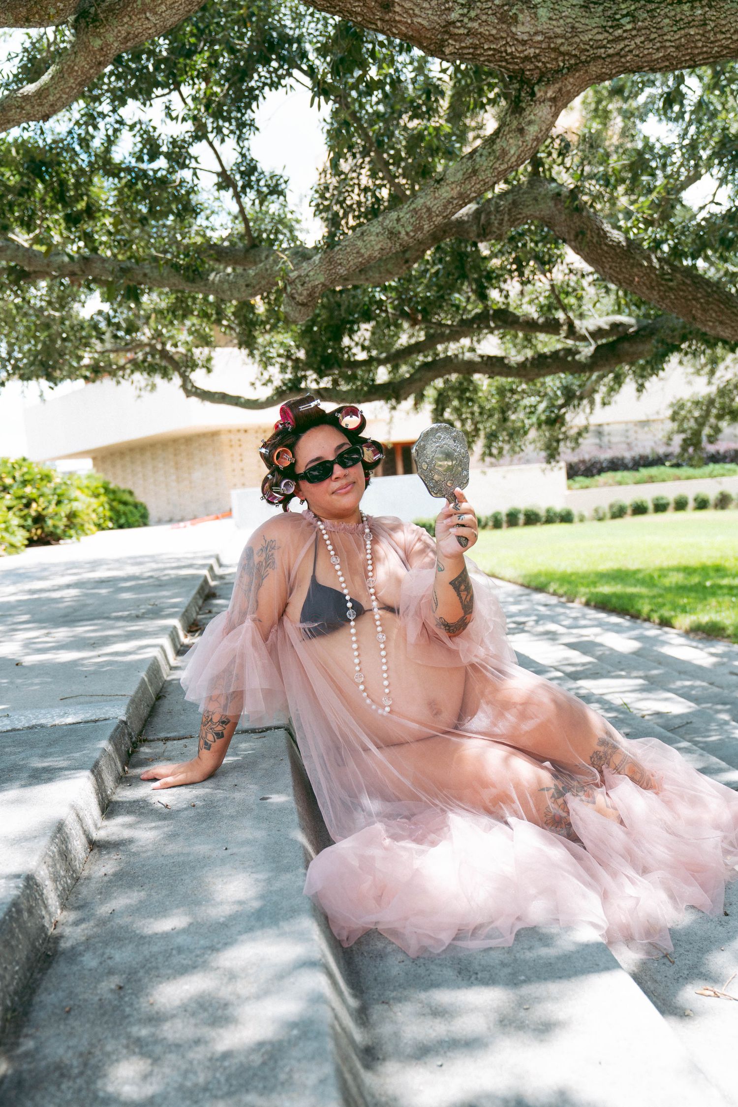 Woman in pink tulle robe, hair curlers, sunglasses, and pearls poses on stone steps holding a mirror outdoors.