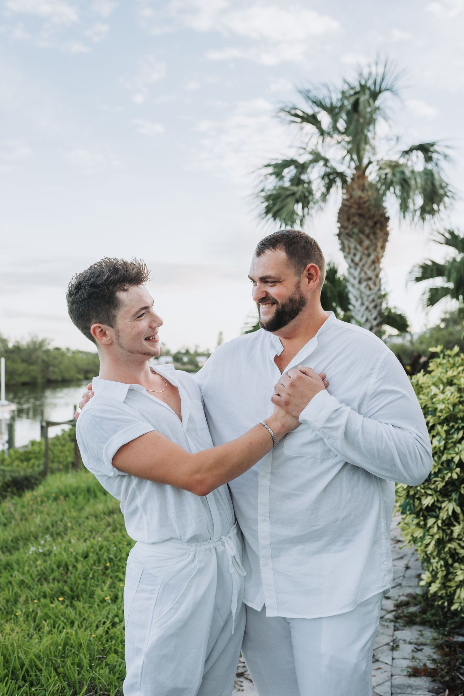 Wedding couple in white clothes sharing intimate moment by palm trees near water.