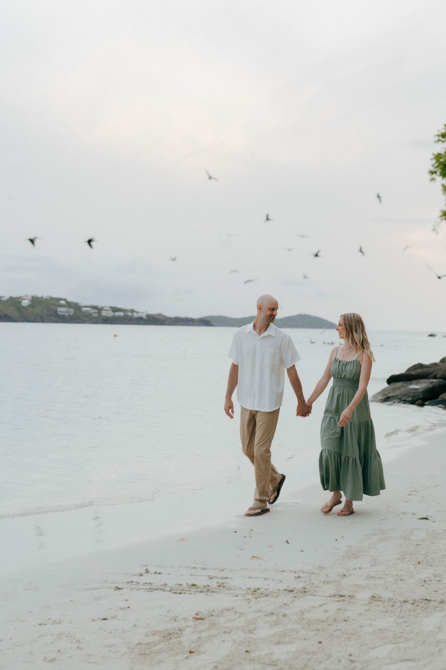 Couple holding hands walking on a white sand beach with birds flying overhead and calm turquoise water in the background.