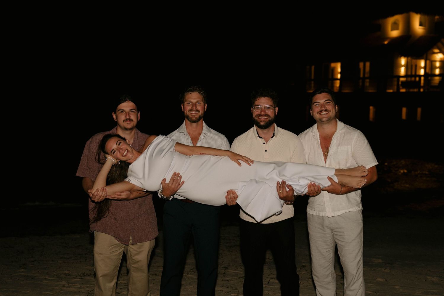 Four men playfully carrying a woman on a beach at night with lit building behind them.