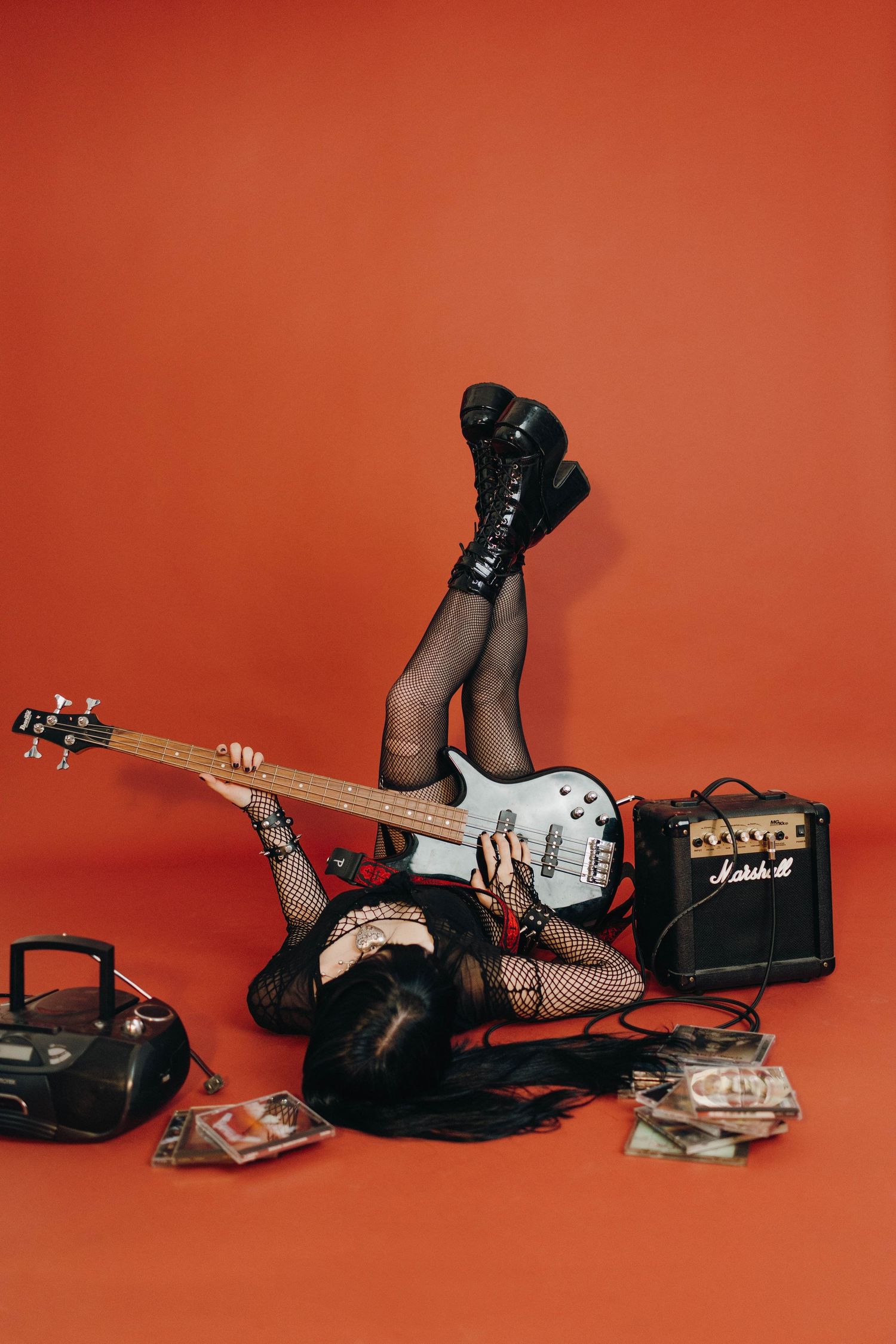 Rock musician lies on red floor with white electric guitar, Marshall amp, boombox, and scattered CDs wearing fishnet tights.