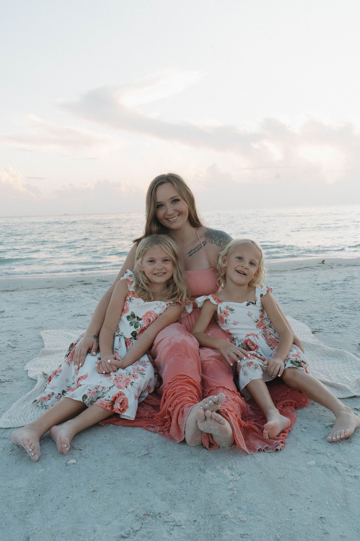 A woman and two young girls in matching floral dresses sit together on a sandy beach at sunset.