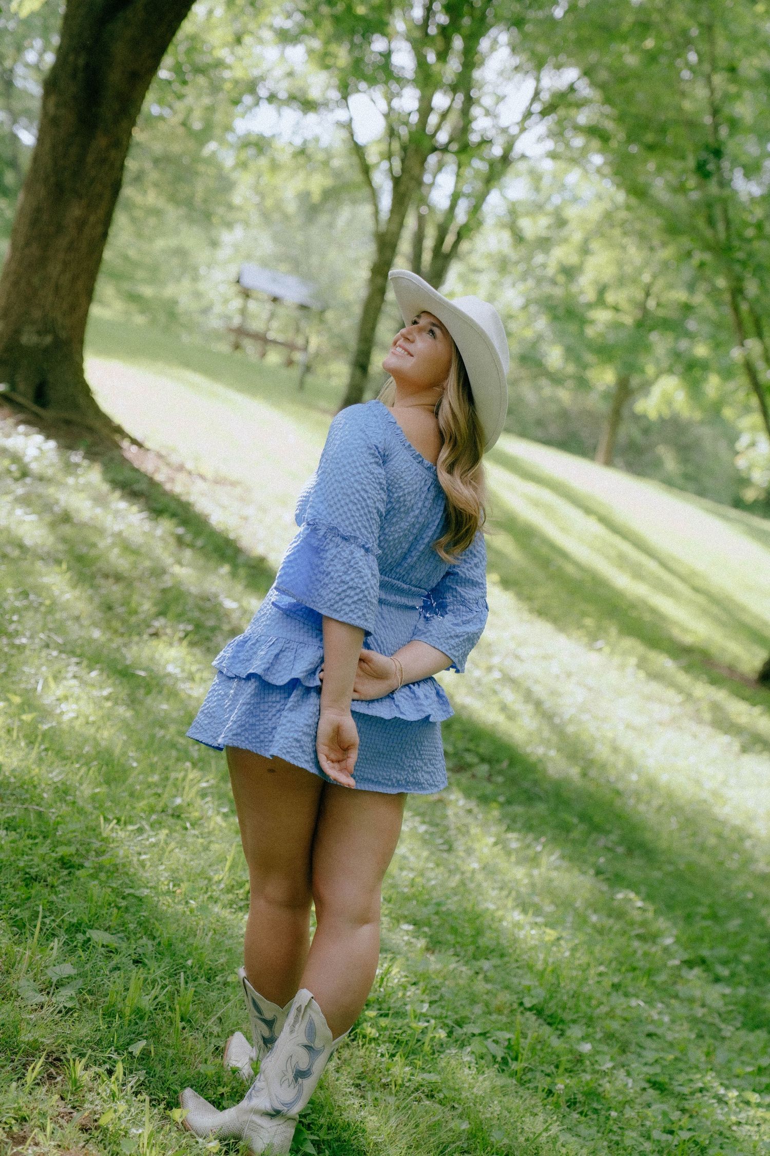 Woman in blue ruffle dress, cowboy boots, and white hat posing outdoors in a lush green park setting.
