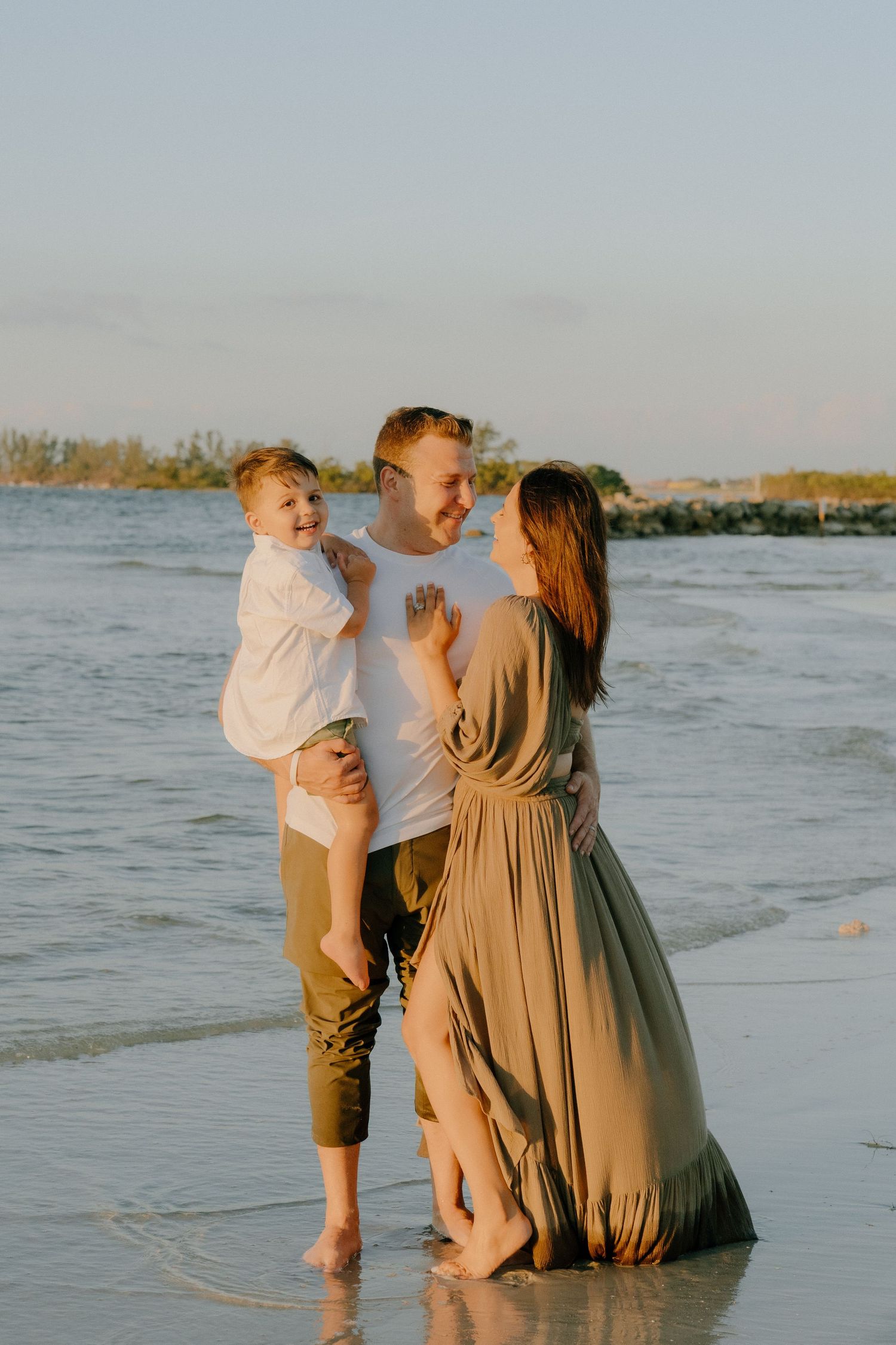 A family stands together at the beach during golden hour, wearing light colored clothing while enjoying the sunset.