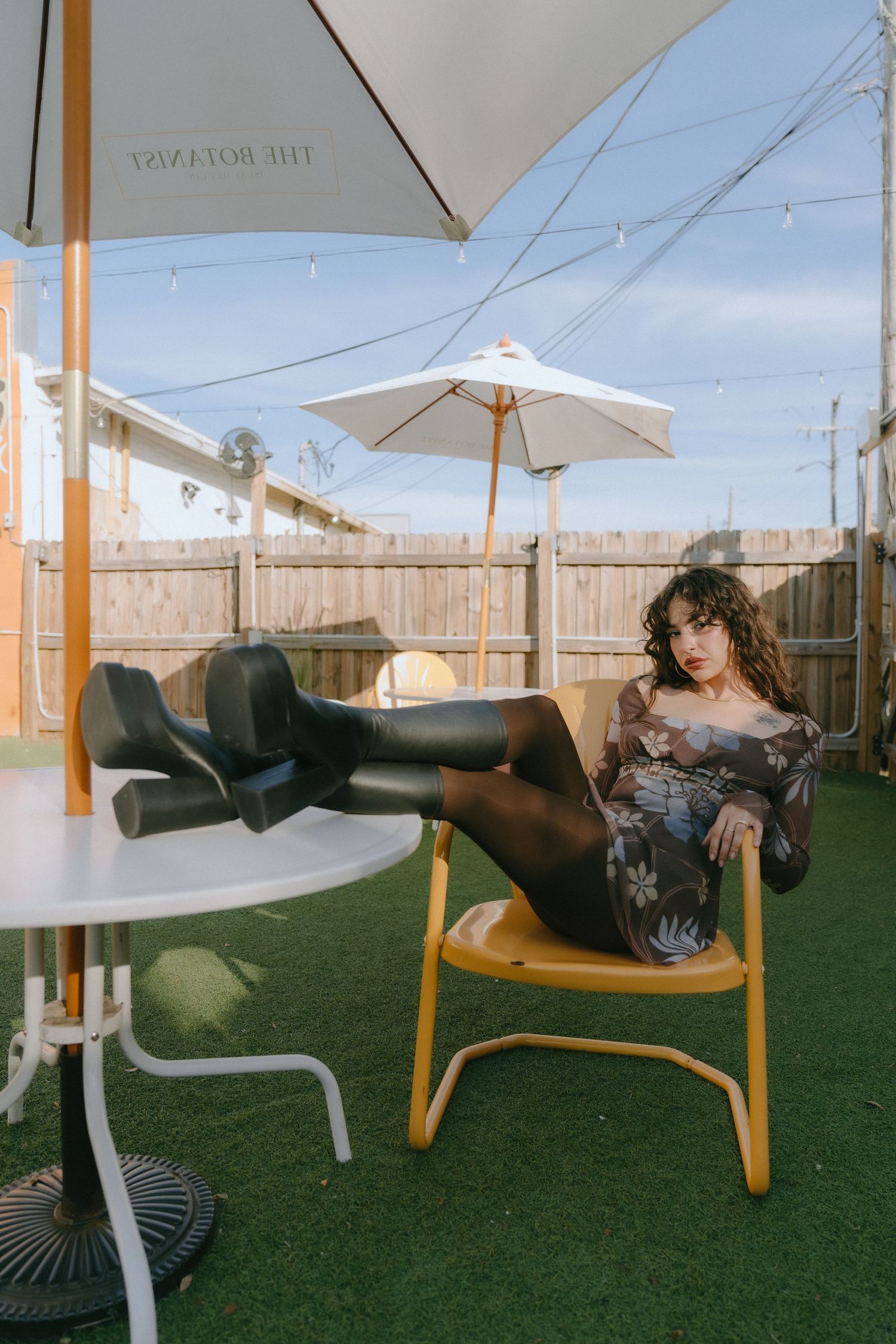Woman in black boots and tights poses on yellow chair in sunny patio with white umbrellas and string lights.