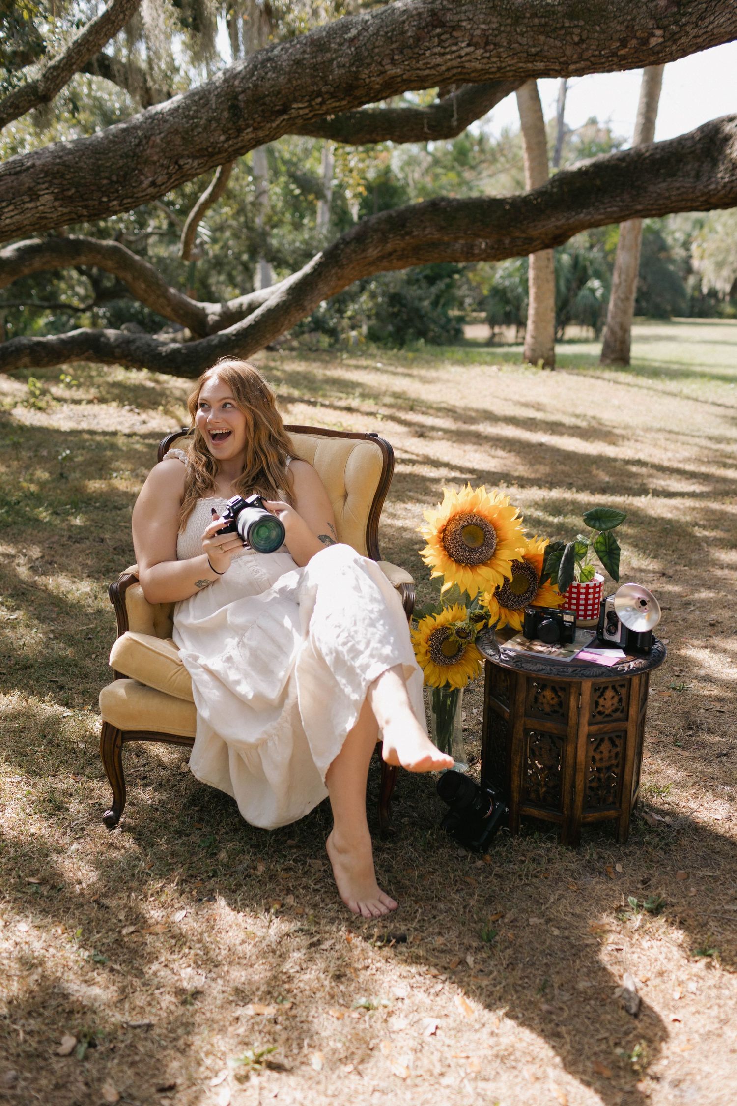 Laughing bride in white dress sits in vintage armchair outdoors, holding camera near sunflowers on ornate side table.
