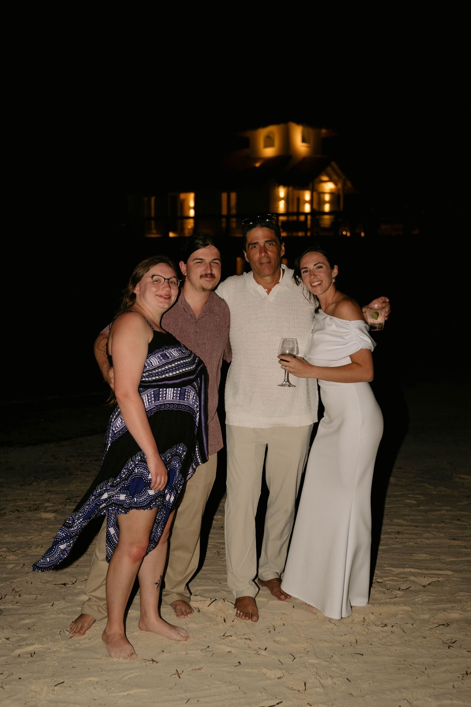 Four people pose on a sandy beach at night with an illuminated building in the background.