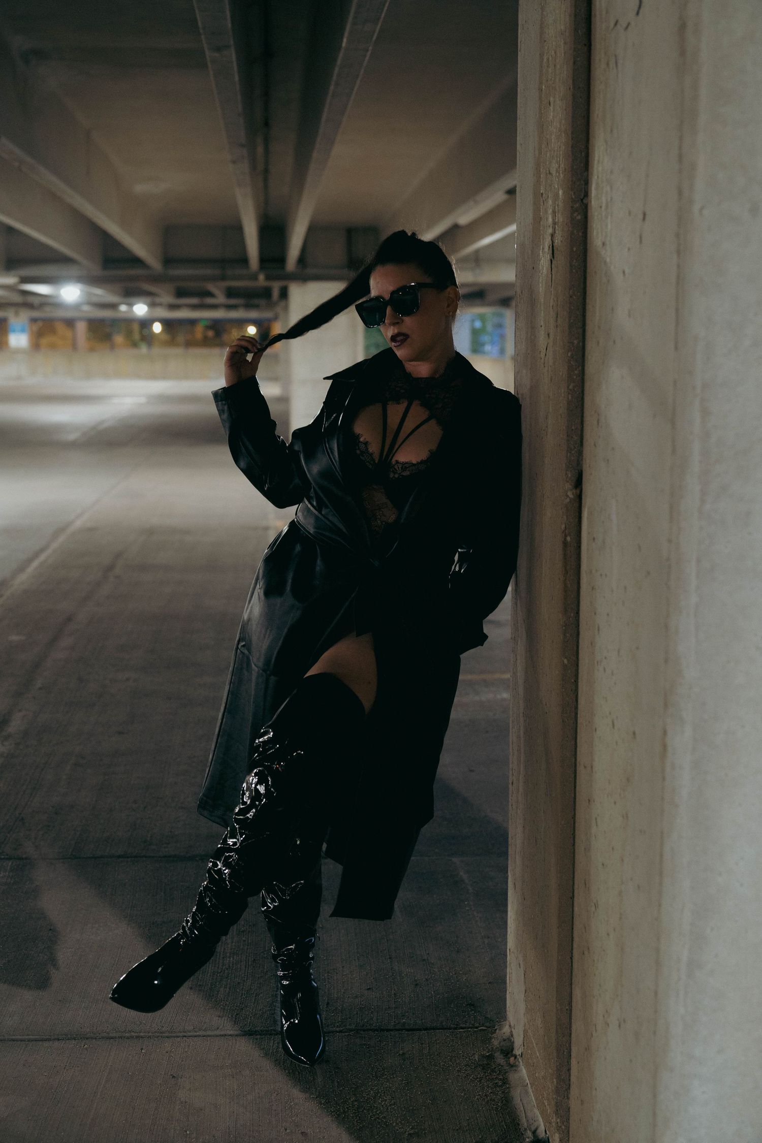 Woman in black trench coat, boots, and sunglasses posing in dimly lit parking garage.